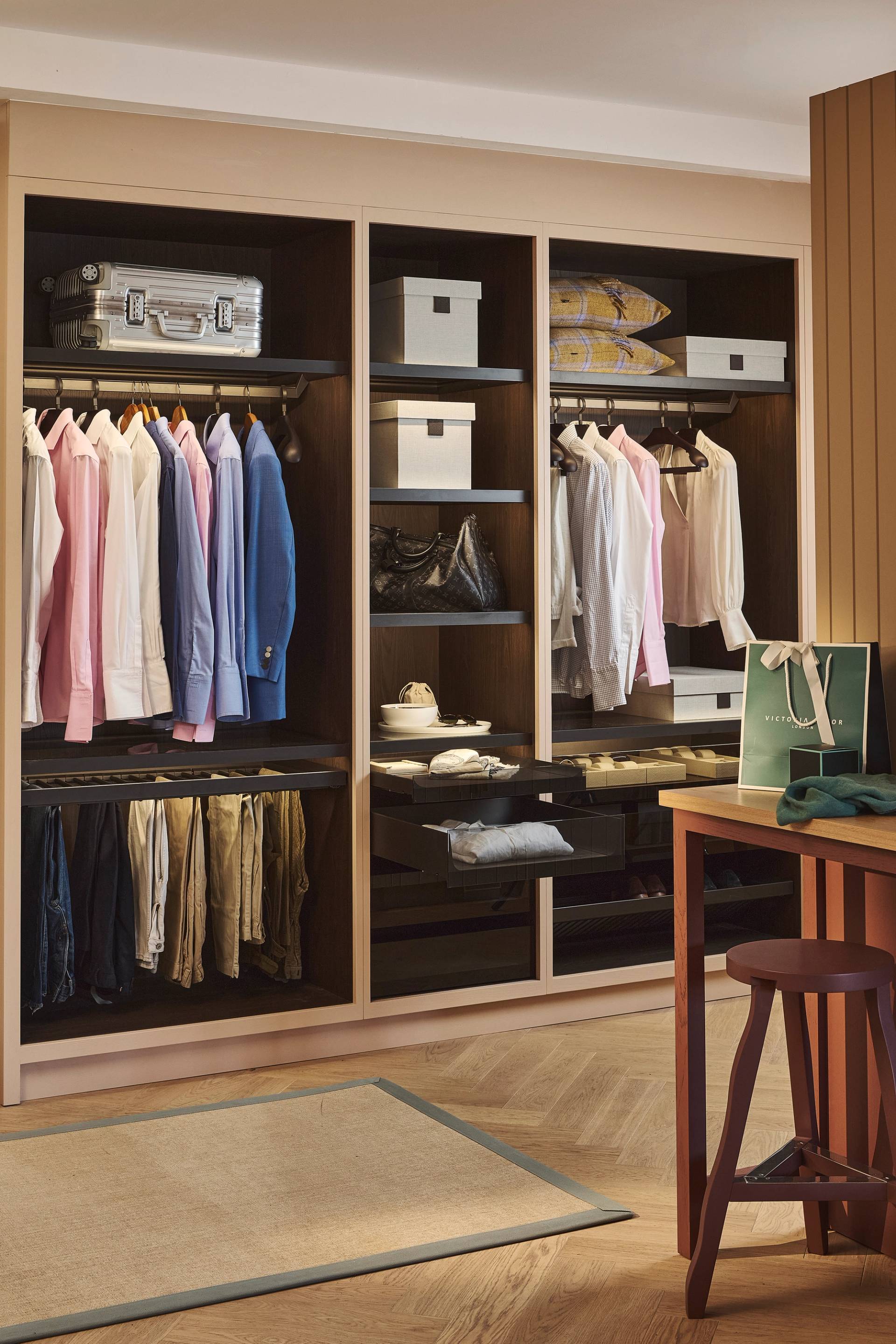 A neatly organized closet with shirts, jackets, and pants hanging, shelves with boxes and bags, folded clothes, and a suitcase. A wooden stool and shopping bag are visible in the foreground.