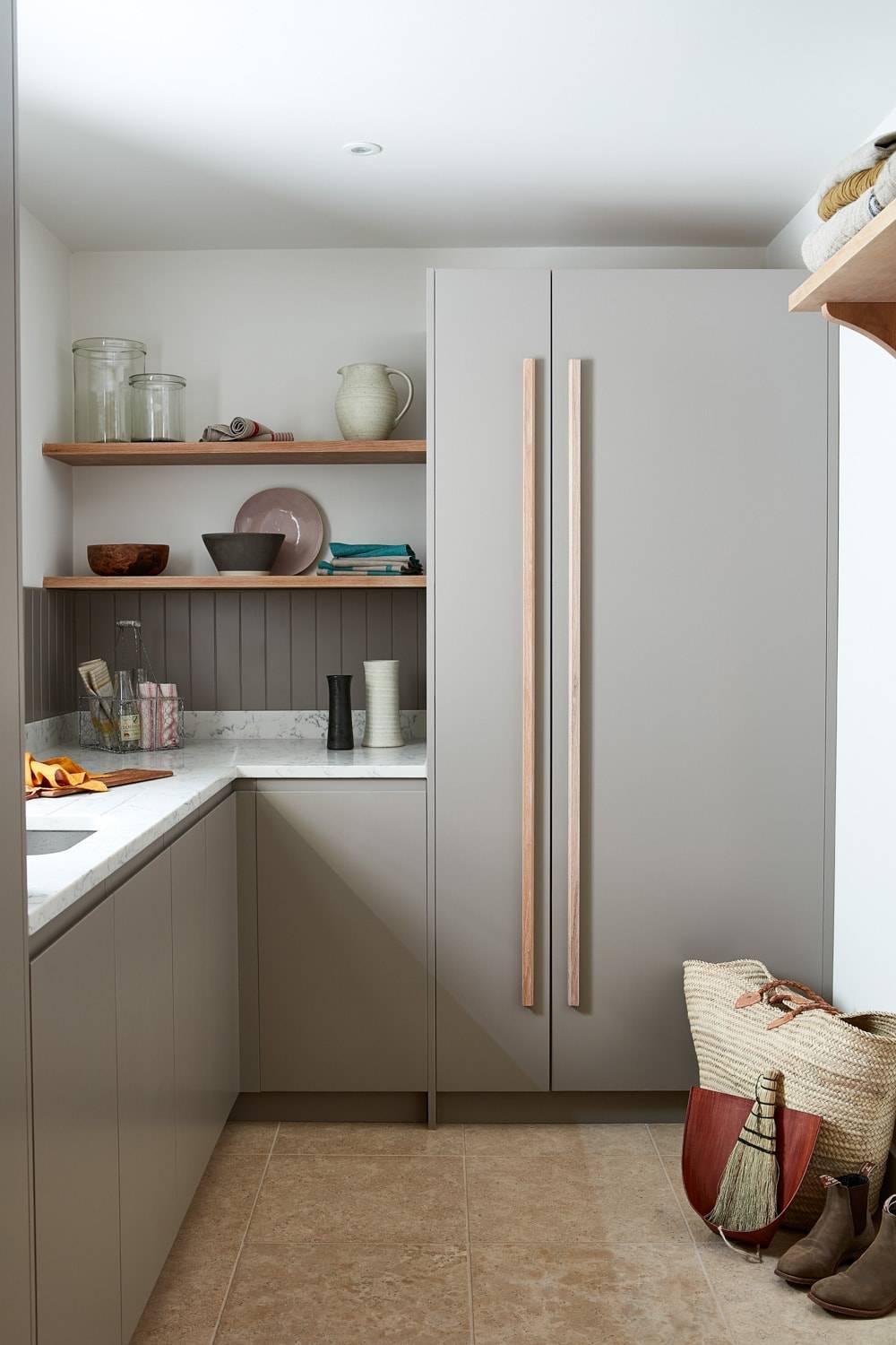 A modern kitchen with light gray cabinets, a large double-door refrigerator, and marble countertops. Open shelves hold various kitchen items. Natural light illuminates the space, showing a tiled floor and a woven basket in the corner.