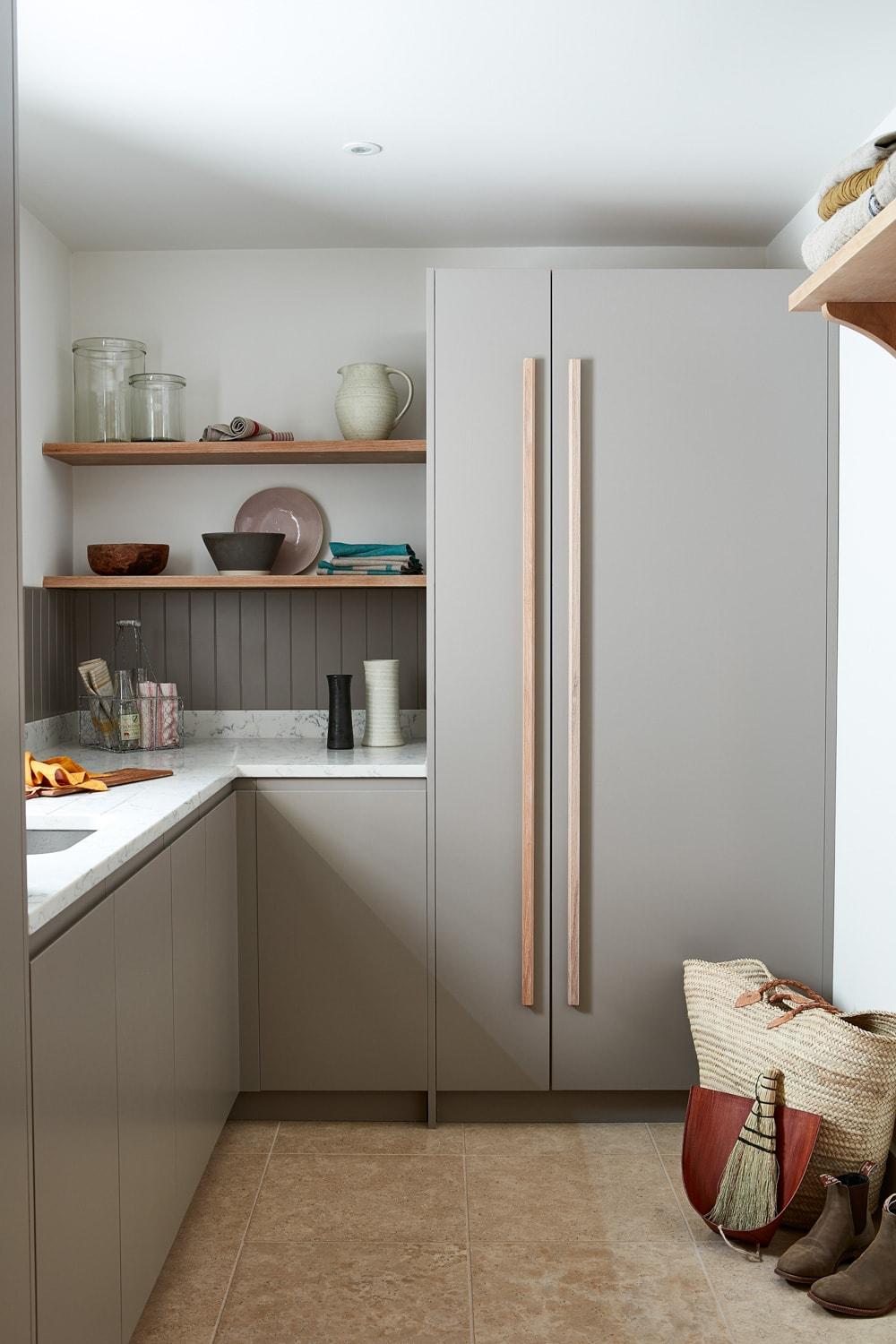 A modern kitchen with light gray cabinets, a large double-door refrigerator, and marble countertops. Open shelves hold various kitchen items. Natural light illuminates the space, showing a tiled floor and a woven basket in the corner.