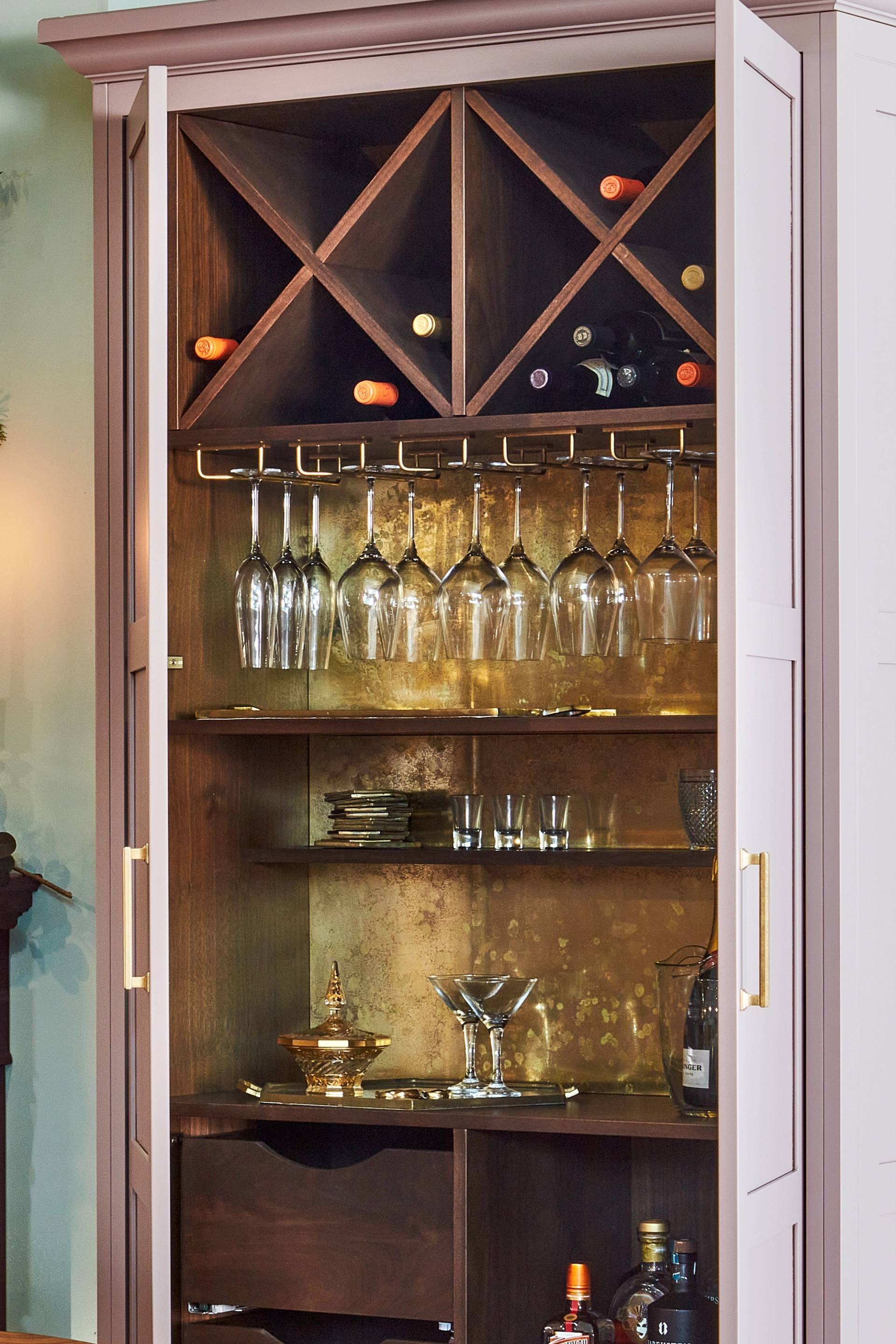 A stylish home bar cabinet with wine racks holding several bottles at the top, glass hangers with various stemware below, and shelves containing glasses, a cocktail shaker, and bottles of liquor. The cabinet has wooden doors with gold handles.
