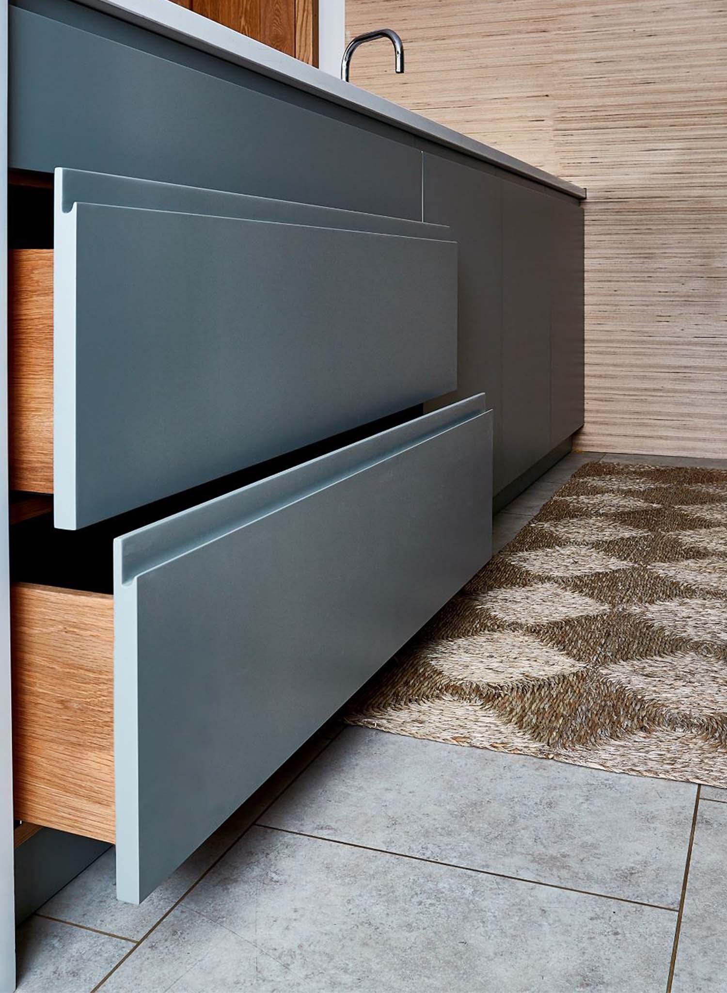 A modern kitchen features open light gray drawers with wooden interiors, a patterned beige rug on the floor, and light-colored tile flooring beside a textured wall and countertop with a sink.