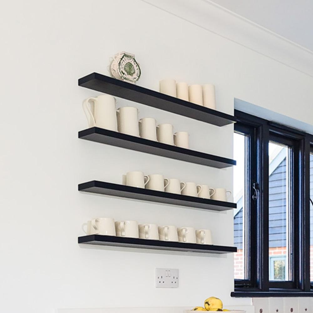 Four black floating shelves on a white wall display white mugs, cups, and a teapot. A window with black trim is on the right side, and there are two lemons on the counter below the shelves.