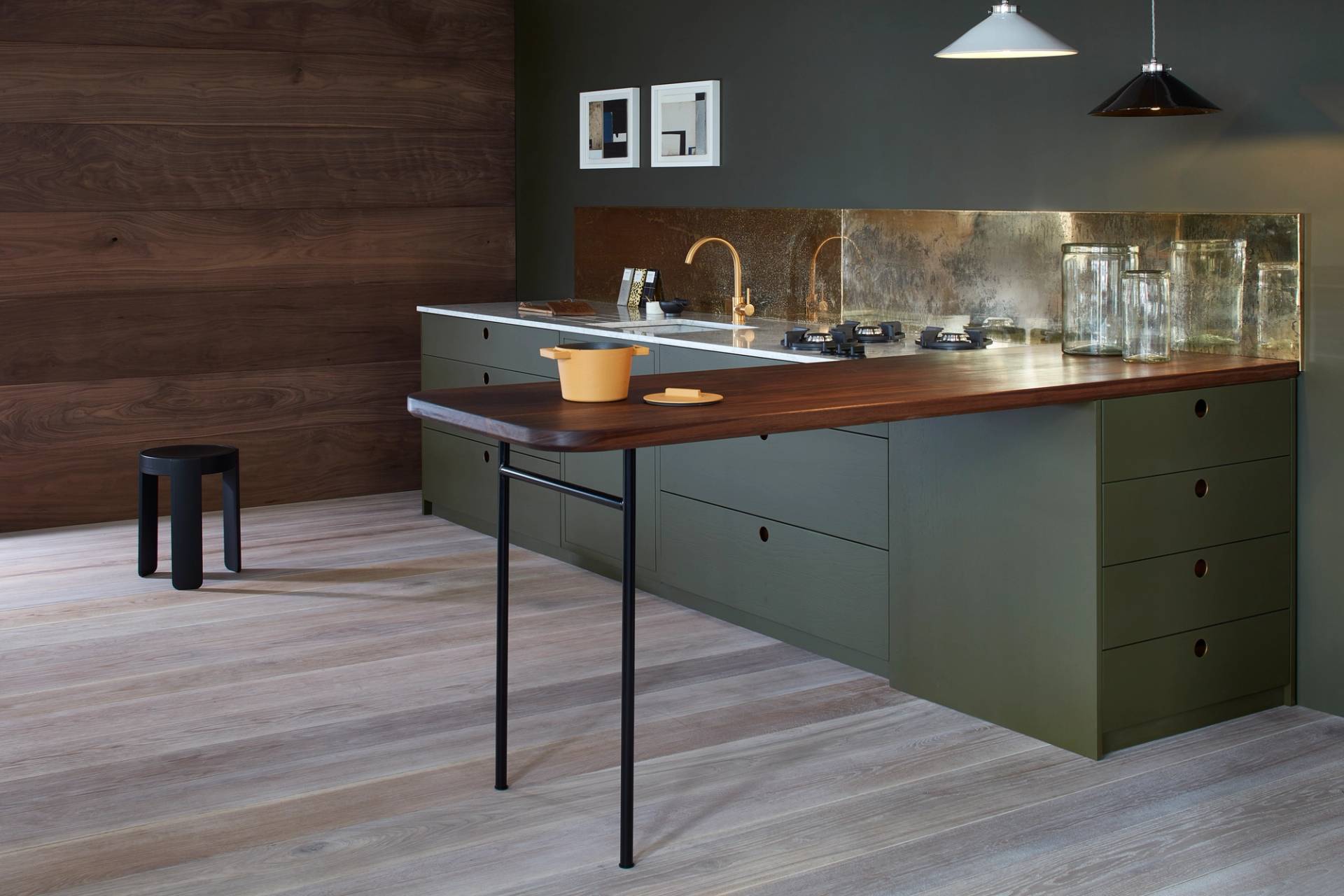 Modern kitchen with green cabinets, brass fixtures, and a wooden countertop that extends as a table with a single black leg. Two pendant lights hang above, and a yellow pot sits on the counter. Walls are dark wood and gray.