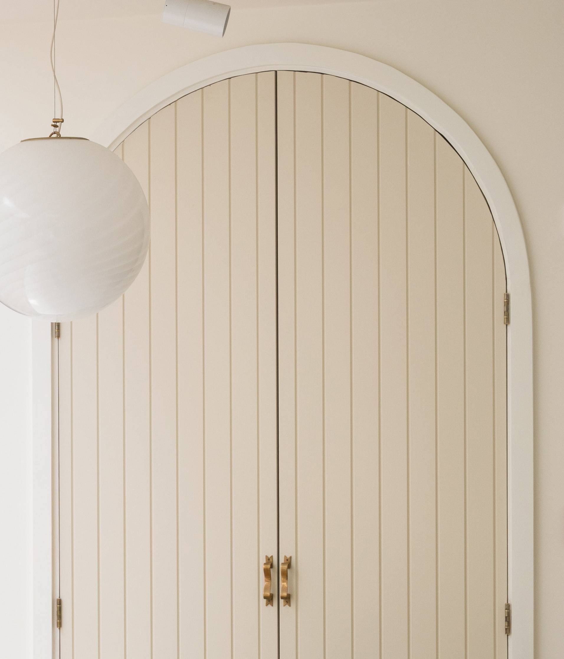 A beige, arched double door with vertical paneling and gold handles stands against a light-colored wall. A round, white pendant light hangs from the ceiling on the left side.