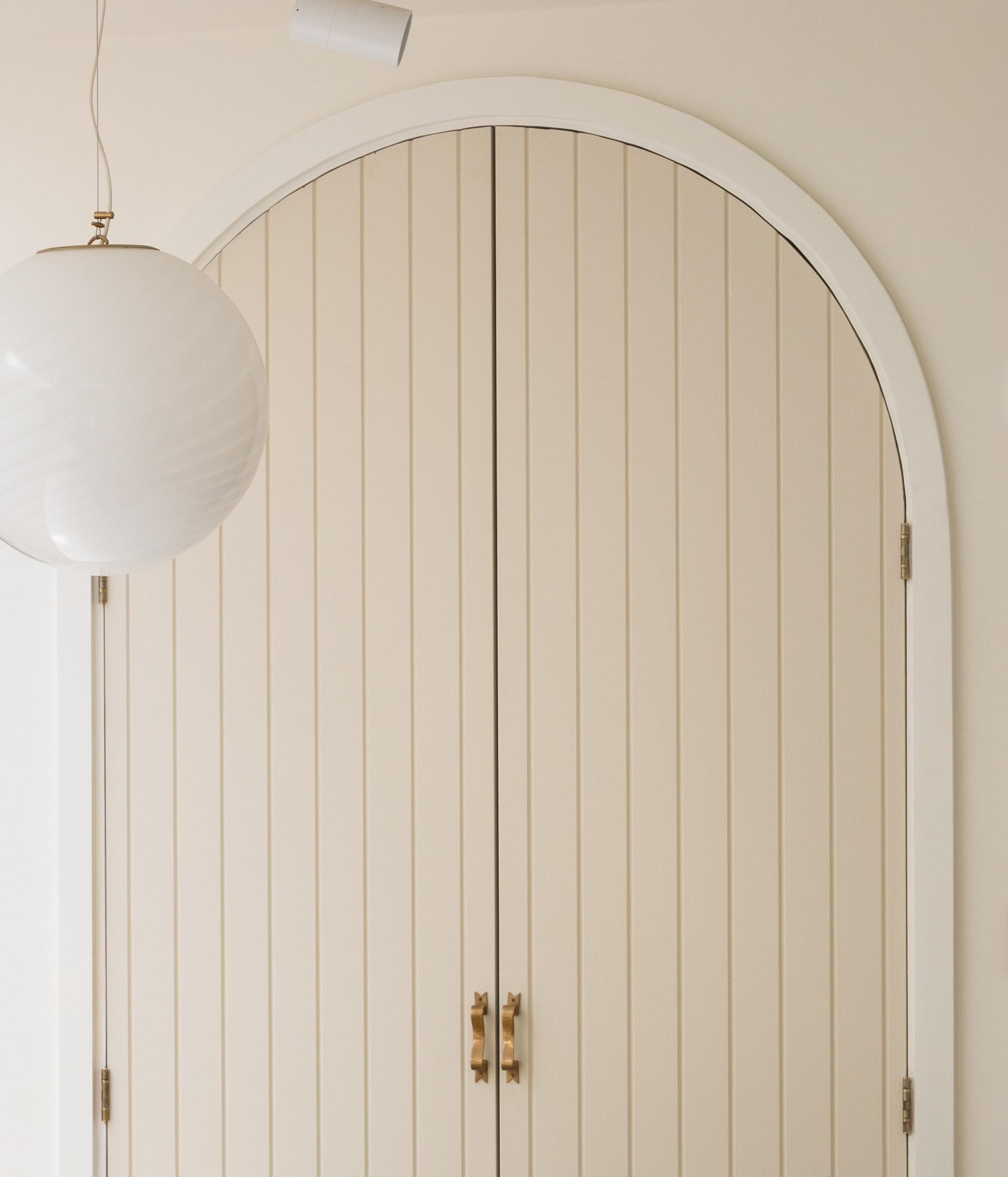 A beige, arched double door with vertical paneling and gold handles stands against a light-colored wall. A round, white pendant light hangs from the ceiling on the left side.
