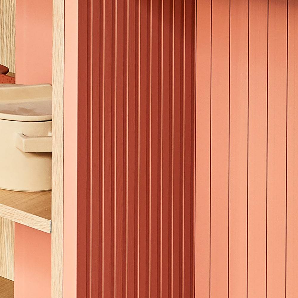 A close-up of a wooden shelf with vertical pink and coral ridged panels. To the left, beige storage containers are partially visible on wooden shelves. The image emphasizes texture and warm, muted colors.
