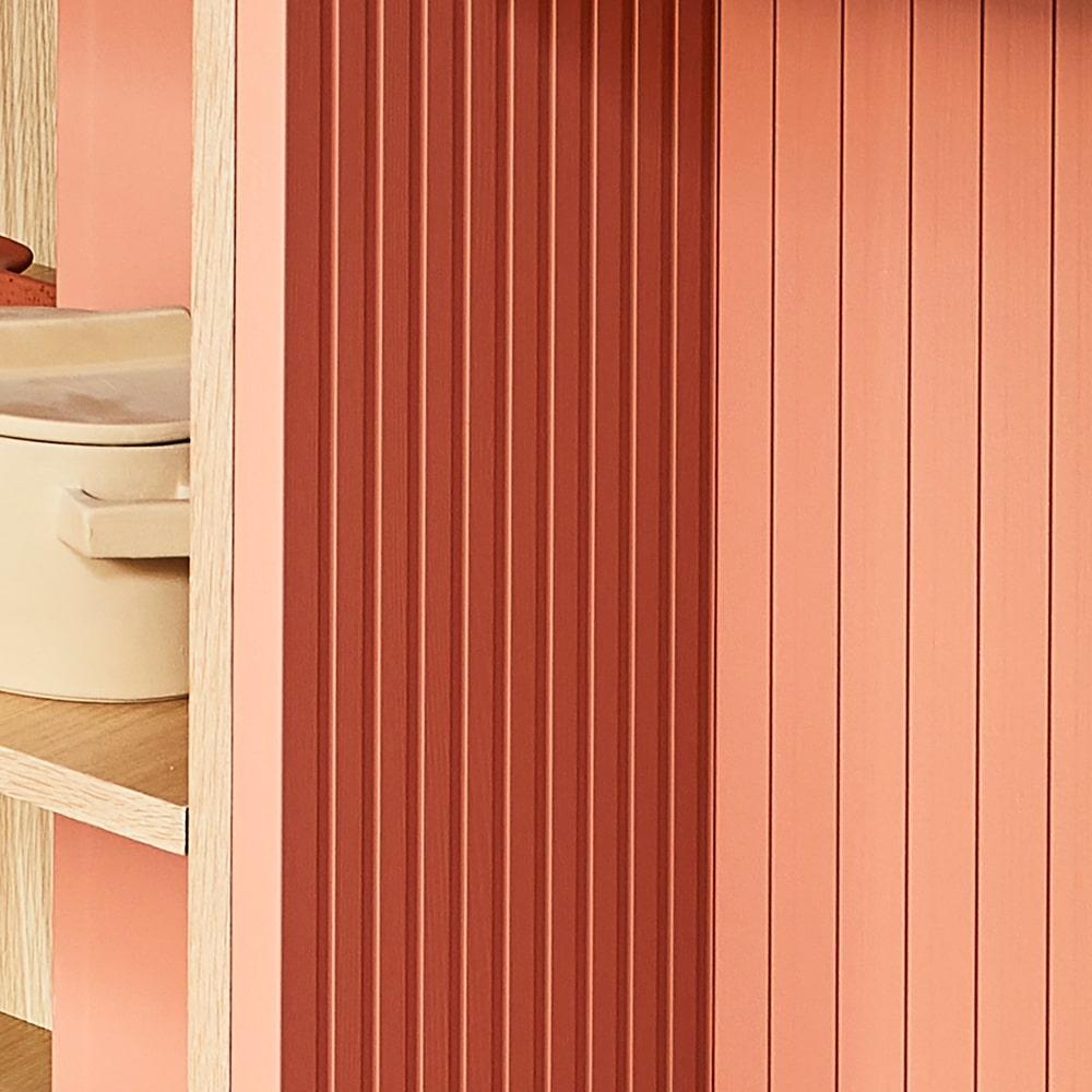 A close-up of a wooden shelf with vertical pink and coral ridged panels. To the left, beige storage containers are partially visible on wooden shelves. The image emphasizes texture and warm, muted colors.