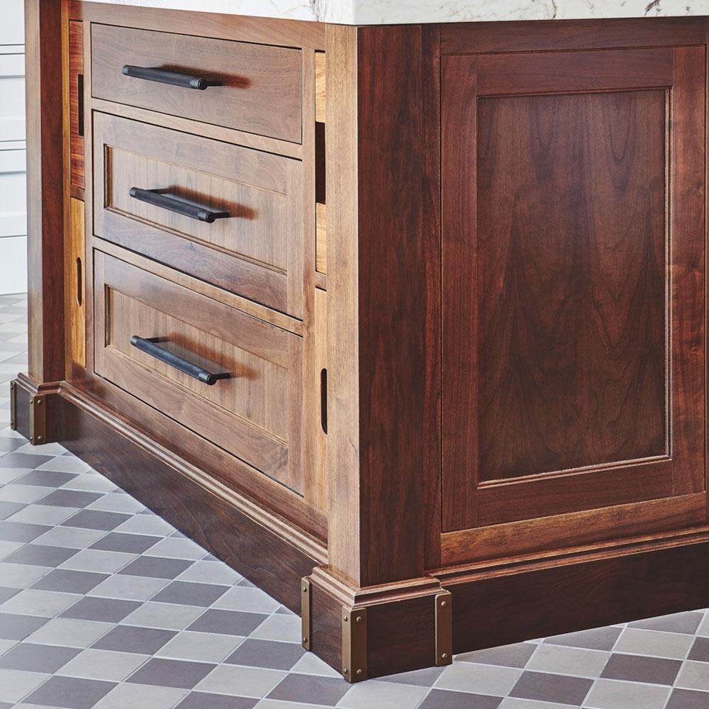 A close-up of a wooden kitchen island with three drawers and black handles, set on a patterned gray and white tiled floor. The island features rich wood grain and detailed trim.