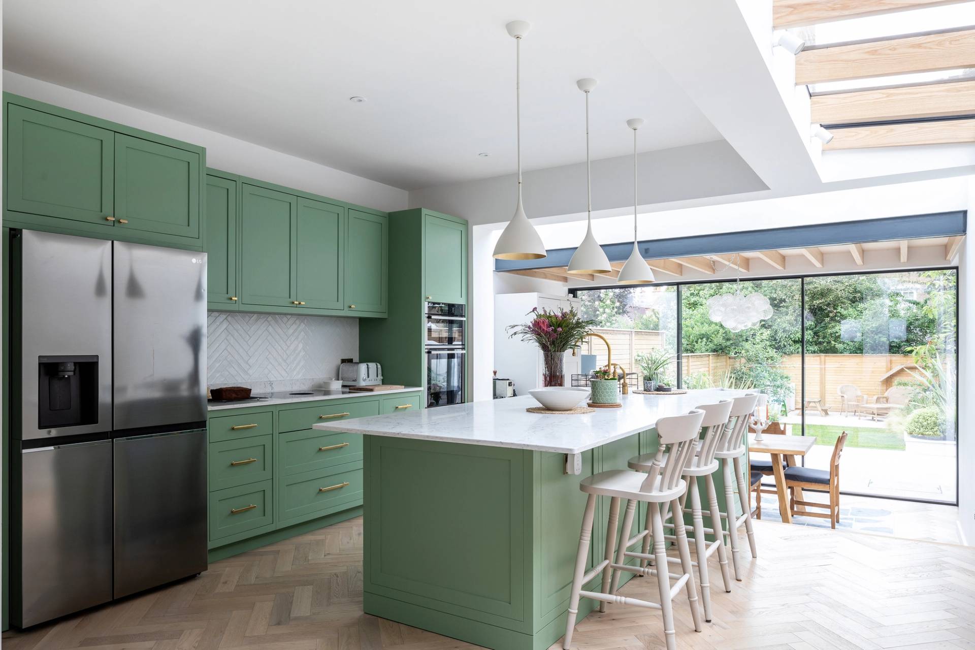 Modern kitchen with green cabinets, white countertops, and a large island with white bar stools. Pendant lights hang above the island. Glass doors open to a sunny garden with a lawn and patio furniture outside.