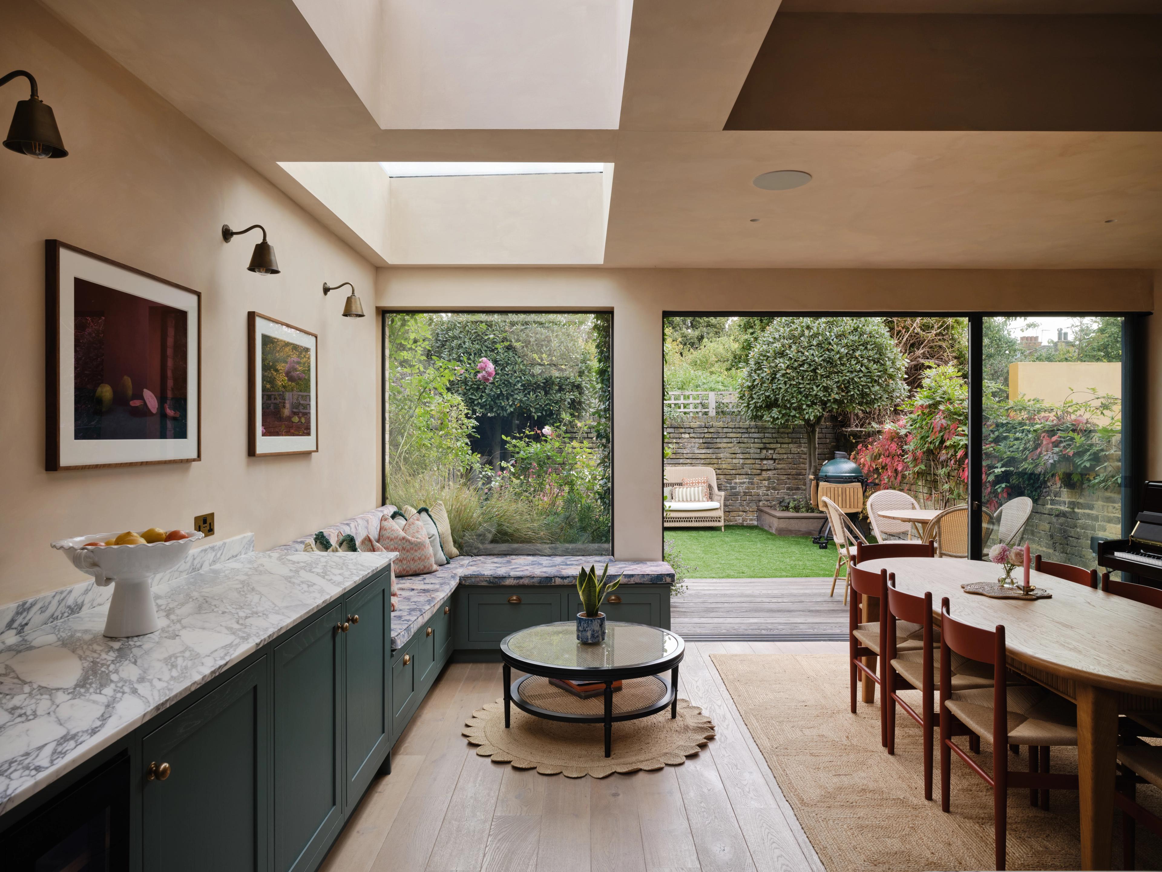 Queens Park kitchen, in-frame Shaker in dark green with a marble worktop, showing an upholstered bench running along the wall beneath a window, a round dining table with wooden chairs in front of it, and open garden doors beyond.
