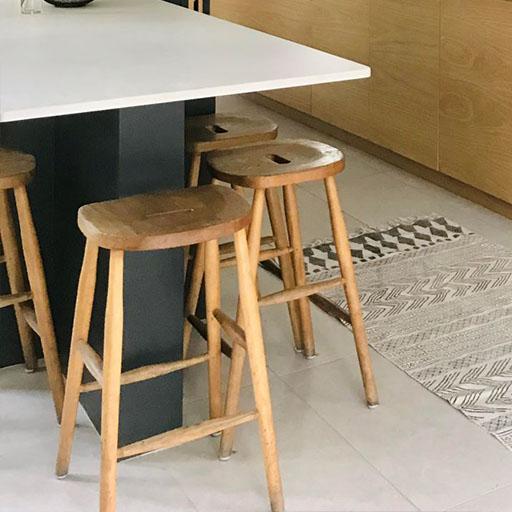 Three wooden bar stools are placed beside a white kitchen island with a black base, near a patterned rug on a light-colored tile floor. Light wood cabinets are visible in the background.