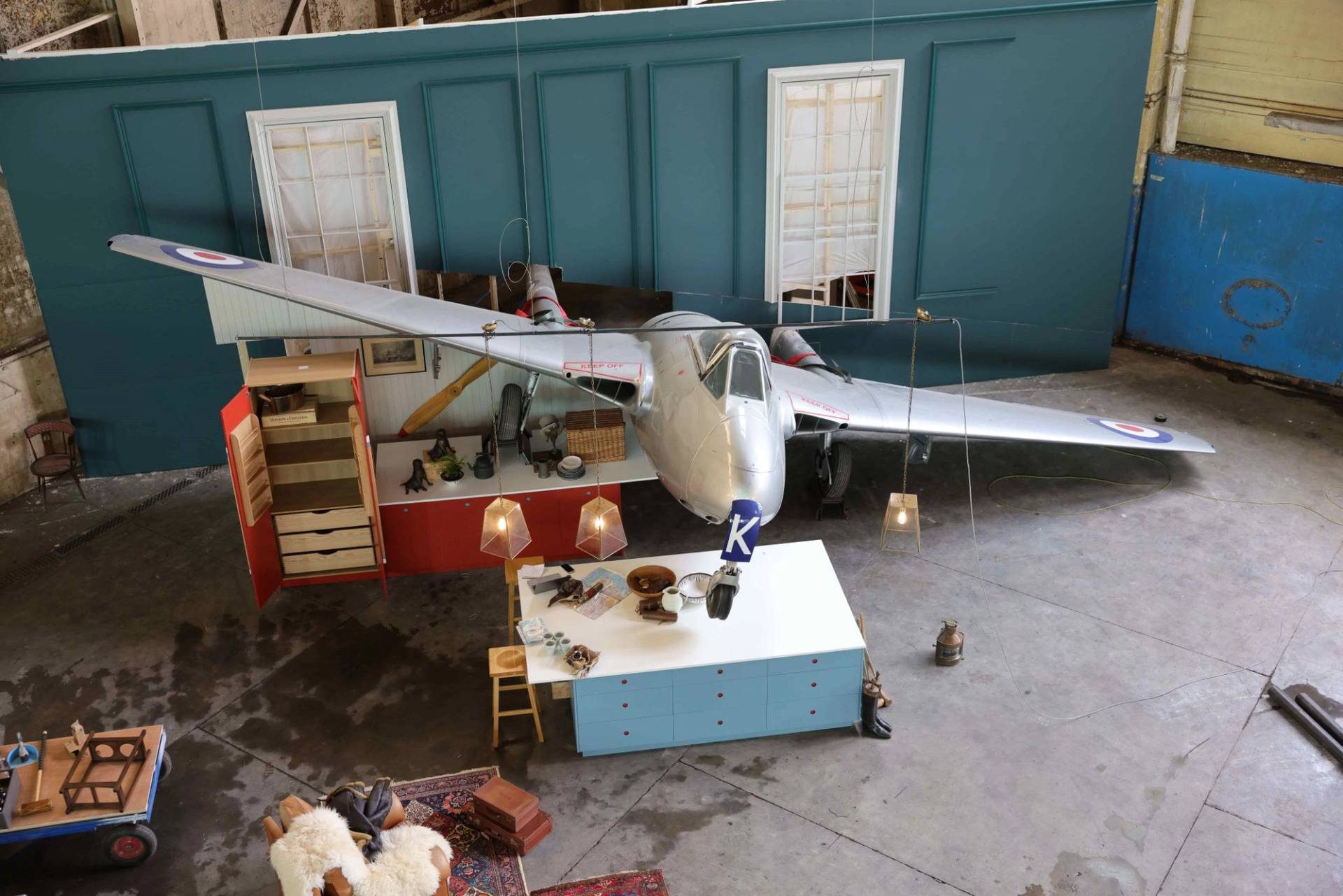 A high-angle, wide shot of an elaborate film or photography set inside a large industrial warehouse. A silver vintage fighter jet with British Royal Air Force roundels is parked in the center, appearing to "crash" through a teal-colored wall. The set is furnished like a quirky living space, featuring a light blue kitchen island, a red cabinet, hanging geometric pendant lights, and various aviation-themed props. The concrete floor and surrounding scaffolding highlight the behind-the-scenes nature of the production.