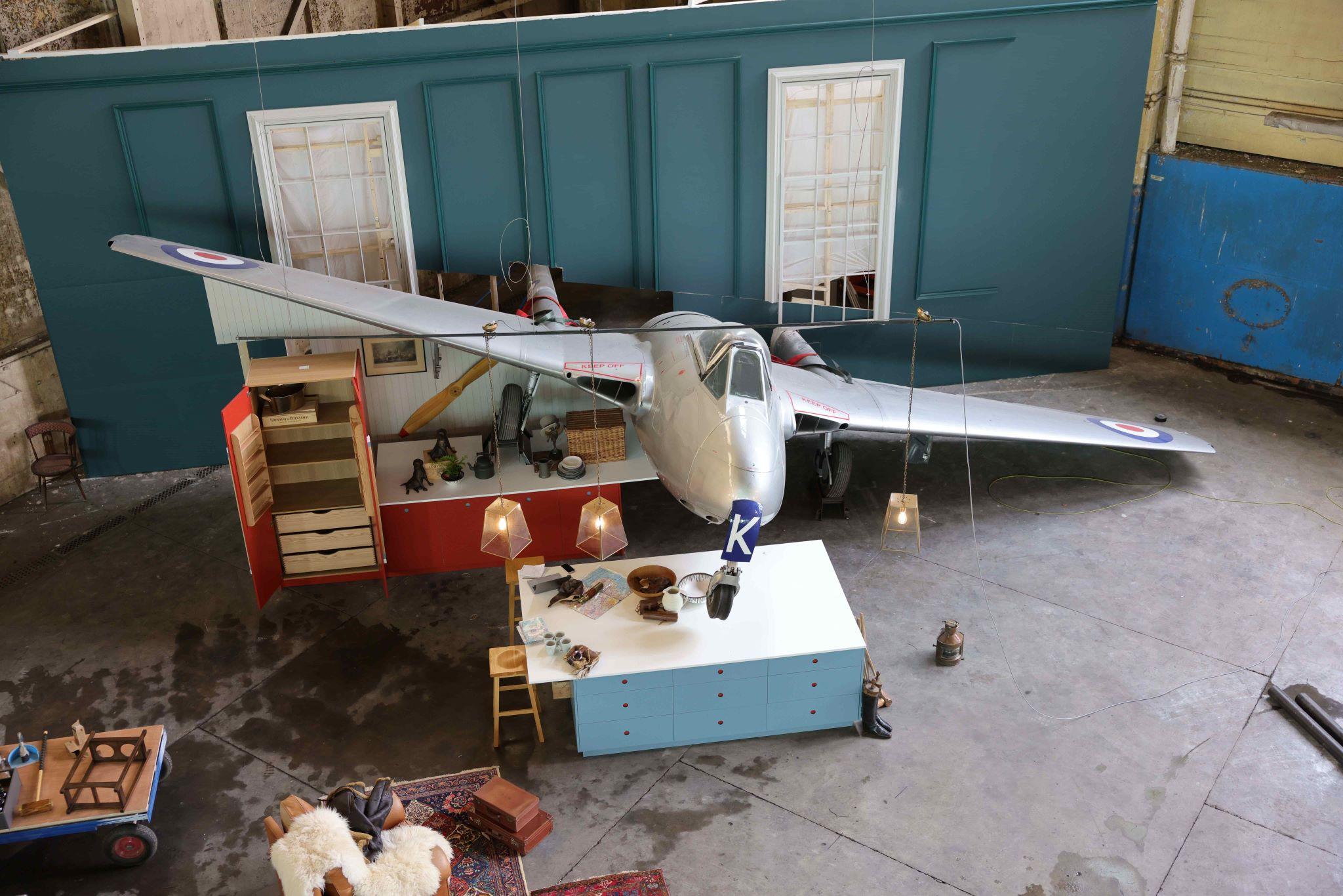 A high-angle, wide shot of an elaborate film or photography set inside a large industrial warehouse. A silver vintage fighter jet with British Royal Air Force roundels is parked in the center, appearing to "crash" through a teal-colored wall. The set is furnished like a quirky living space, featuring a light blue kitchen island, a red cabinet, hanging geometric pendant lights, and various aviation-themed props. The concrete floor and surrounding scaffolding highlight the behind-the-scenes nature of the production.