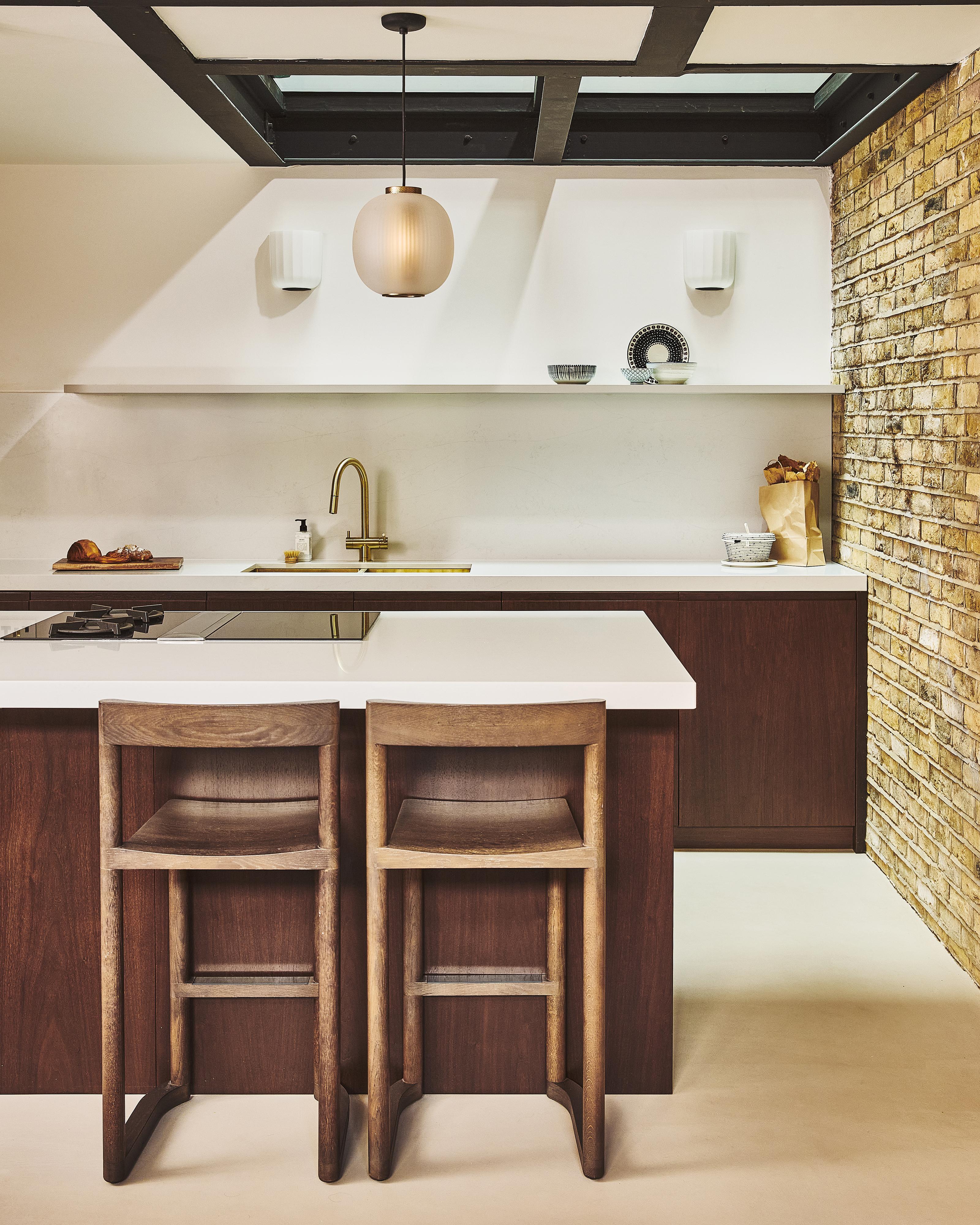Modern kitchen with a central island, two wooden bar stools, white countertops, built-in ovens, minimalist cabinets, pendant lights, and an exposed brick wall on the right.
