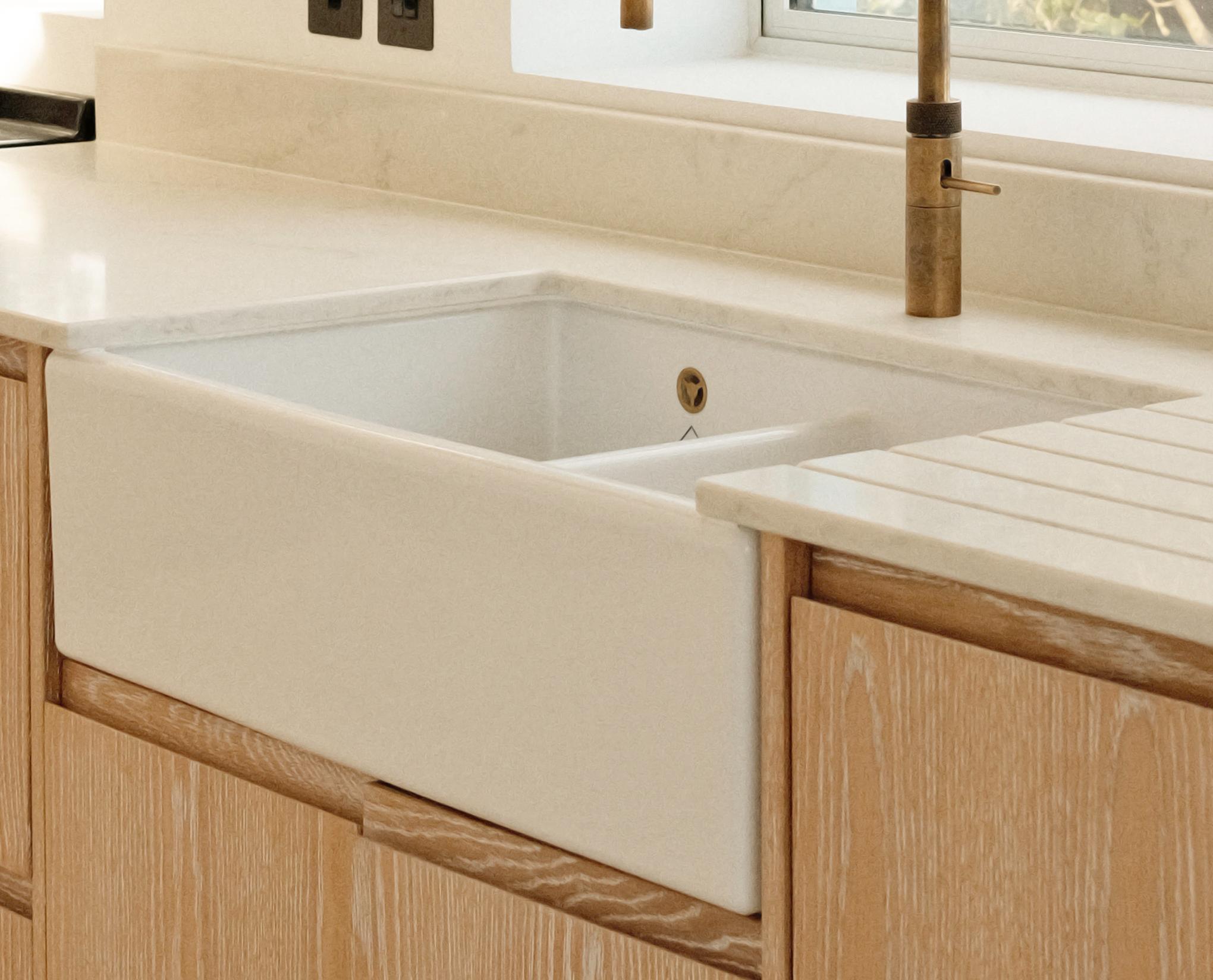 A modern kitchen features a white farmhouse sink with a brass faucet, set in a light wood cabinet with a white countertop, beneath a window allowing natural light into the space.