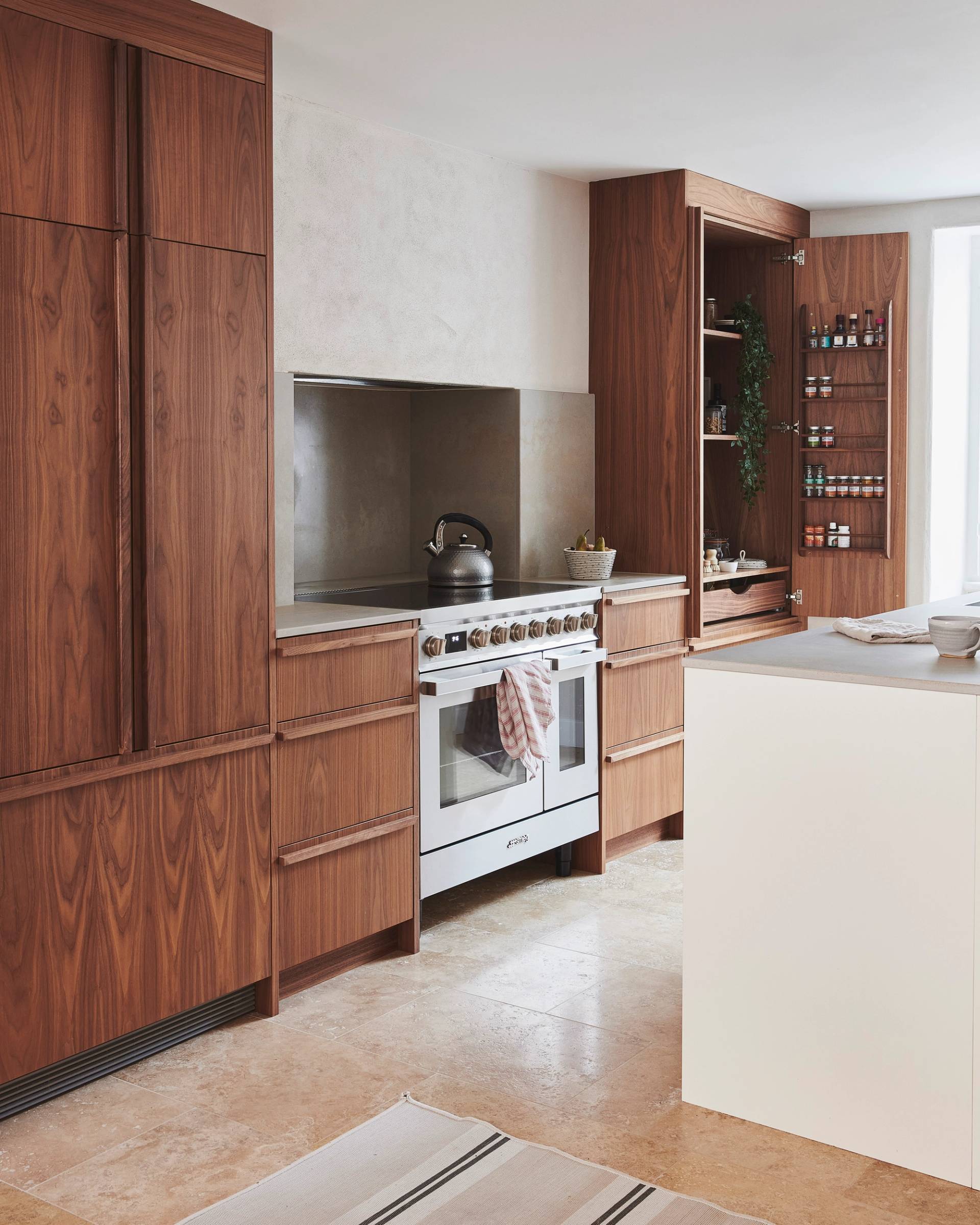 Modern kitchen with wooden cabinets featuring a white oven and stovetop. A kettle sits on the stove. Open shelving with spices and a plant is on the right. A central island with a light countertop is in the foreground, and a striped rug is on the floor.