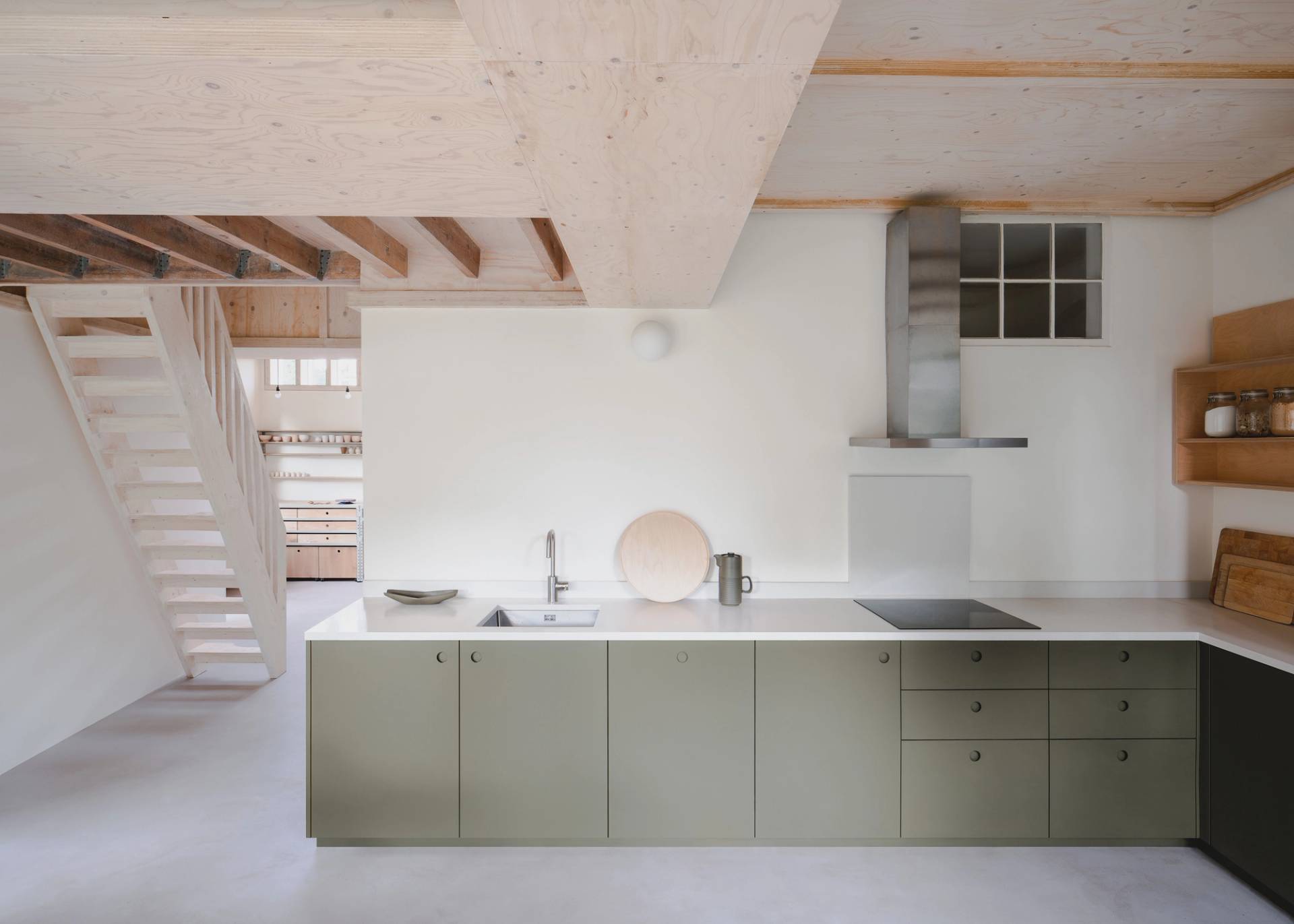 Minimalist kitchen and dining area with light wood furniture, green cabinets, and a large window overlooking greenery. An open staircase leads to the upper floor, and natural light fills the space, highlighting the clean, modern design
