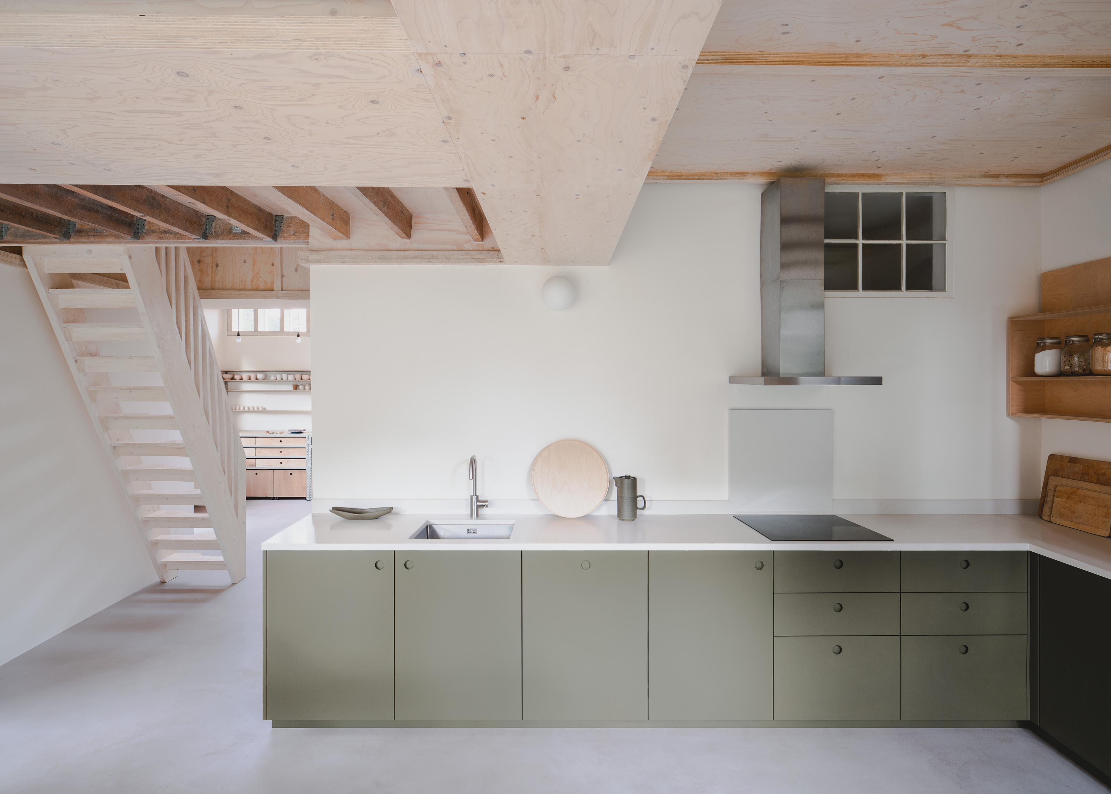 Minimalist kitchen and dining area with light wood furniture, green cabinets, and a large window overlooking greenery. An open staircase leads to the upper floor, and natural light fills the space, highlighting the clean, modern design