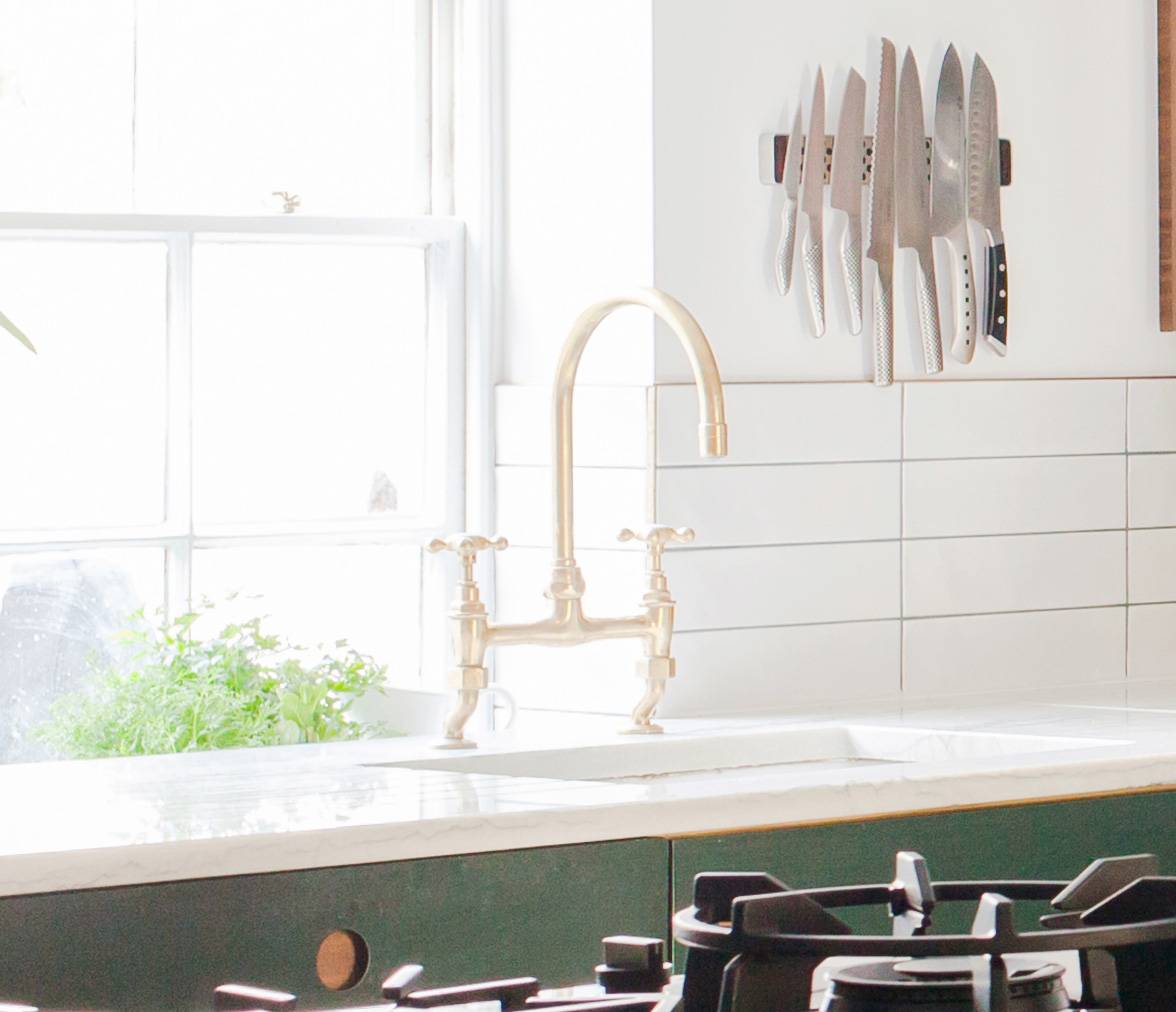 A bright kitchen features a marble countertop with a brass faucet, white tiled backsplash, and a knife holder displaying knives. A gas stove is in the foreground, and a lush green plant sits by a sunlit window.