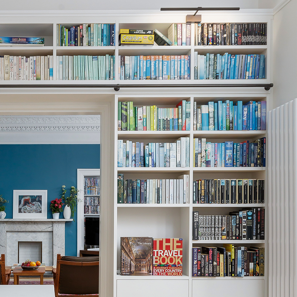 A large built-in bookshelf filled with colorful books, organized mostly by color, stands in a bright room. An open doorway reveals a blue-walled room with a fireplace, a painting, and flower arrangements.