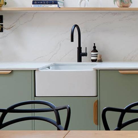 A modern kitchen with green cabinets, a white farmhouse sink, a black faucet, soap dispenser, a scrub brush, and marble backsplash. Open shelves above hold books and a small plant. Black chairs are in the foreground.