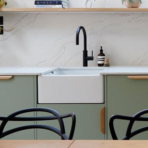 A modern kitchen with green cabinets, a white farmhouse sink, a black faucet, soap dispenser, a scrub brush, and marble backsplash. Open shelves above hold books and a small plant. Black chairs are in the foreground.
