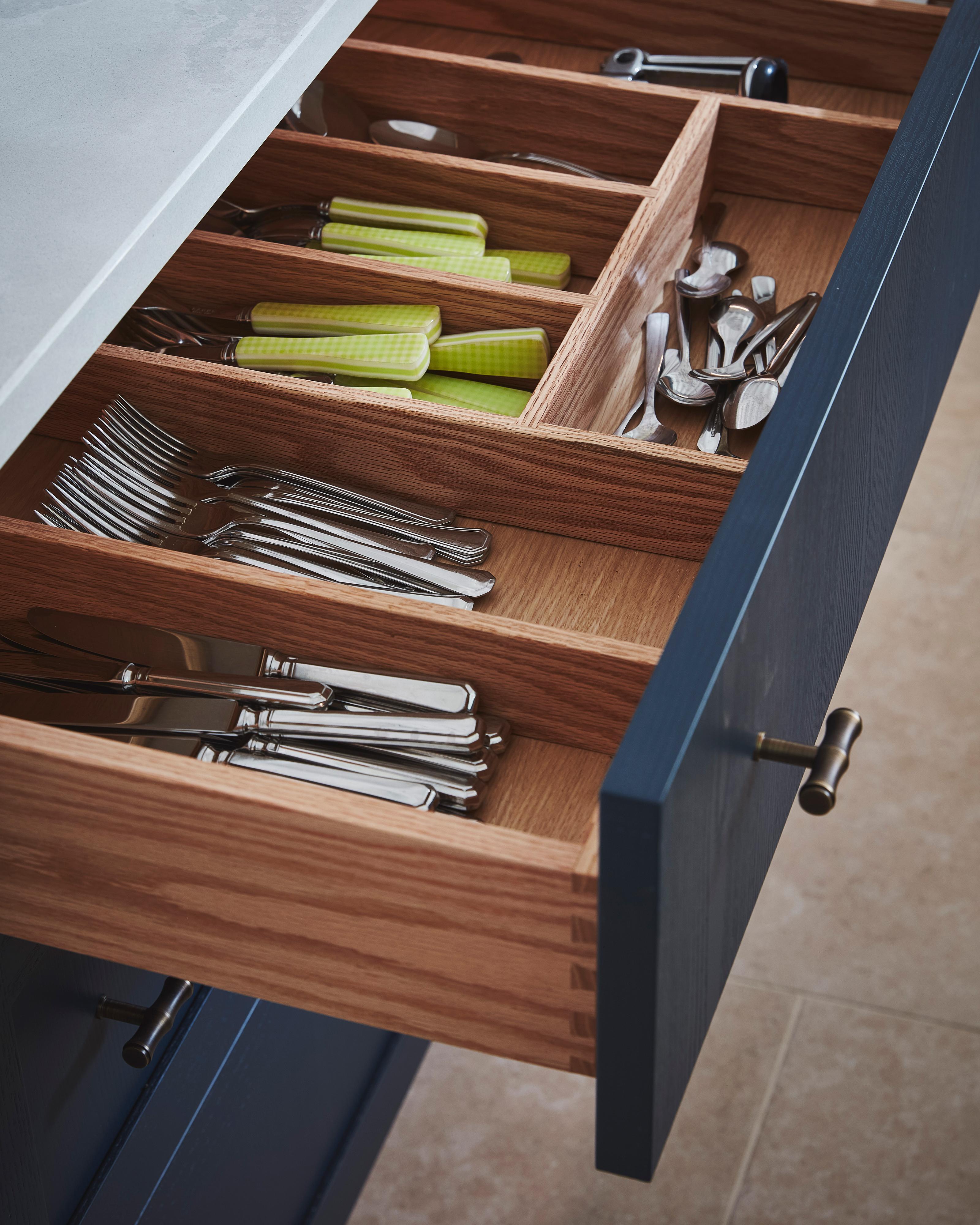 An open wooden kitchen drawer with organized compartments holding silverware—knives, forks, spoons—and a set of green-handled utensils in a navy blue cabinet.