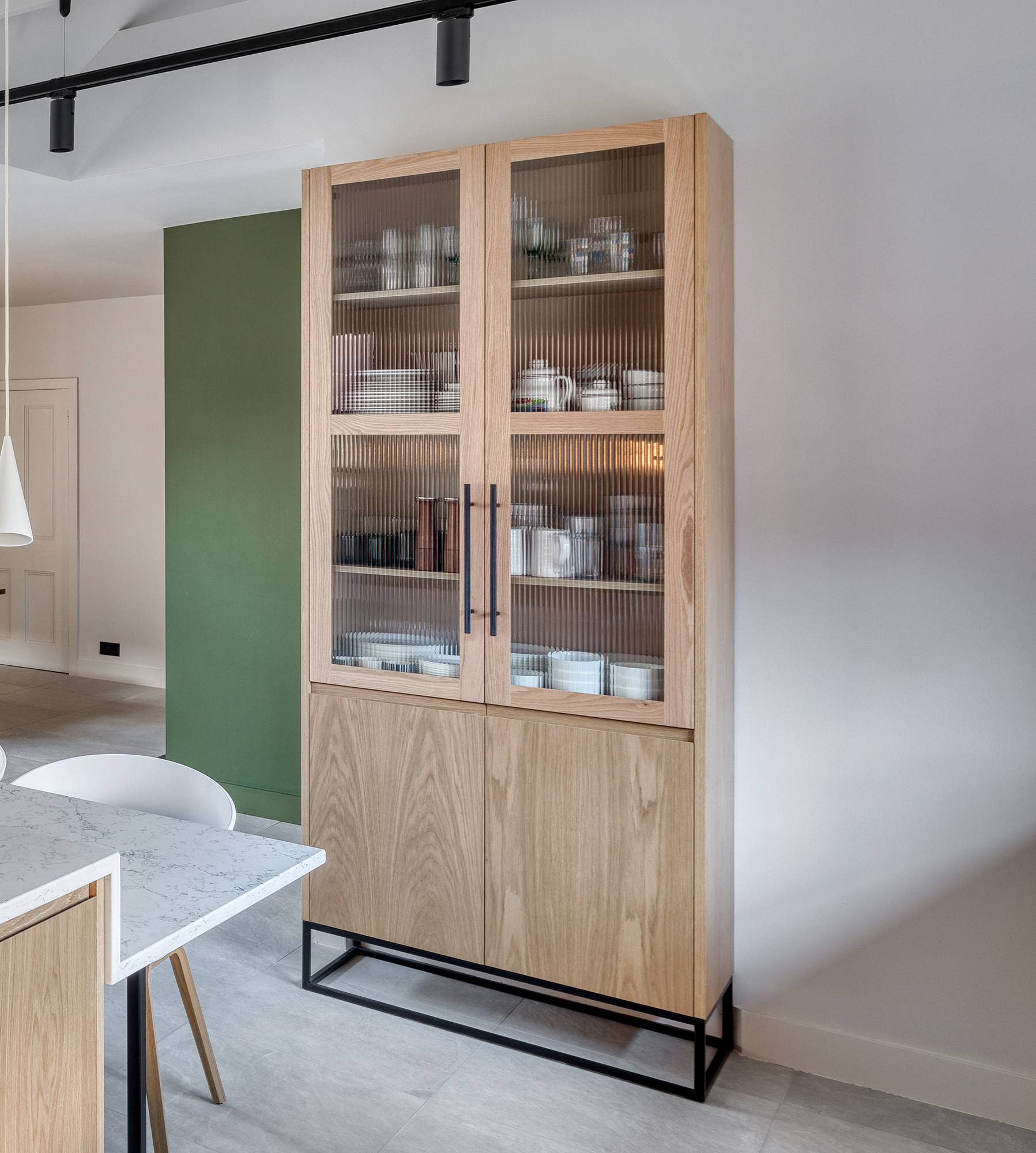 A wooden cabinet with glass doors displays neatly arranged white dishes. It stands in a modern kitchen with light walls and a green accent wall. A pendant light and part of a white marble table are visible in the foreground.