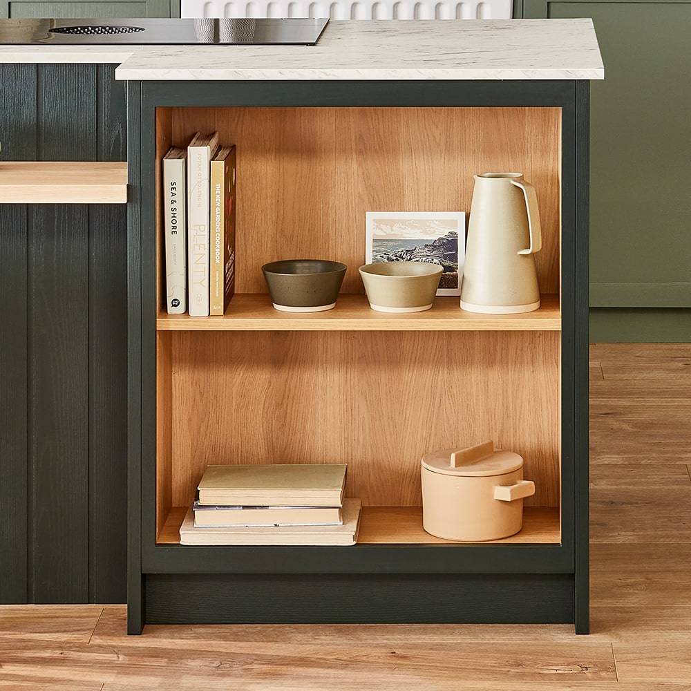 Open wooden kitchen shelf with black sides. Top shelf holds books, several bowls, a small painting, and a cream pitcher. Bottom shelf contains stacked books and a round beige container. Light wooden floor and marble countertop visible.