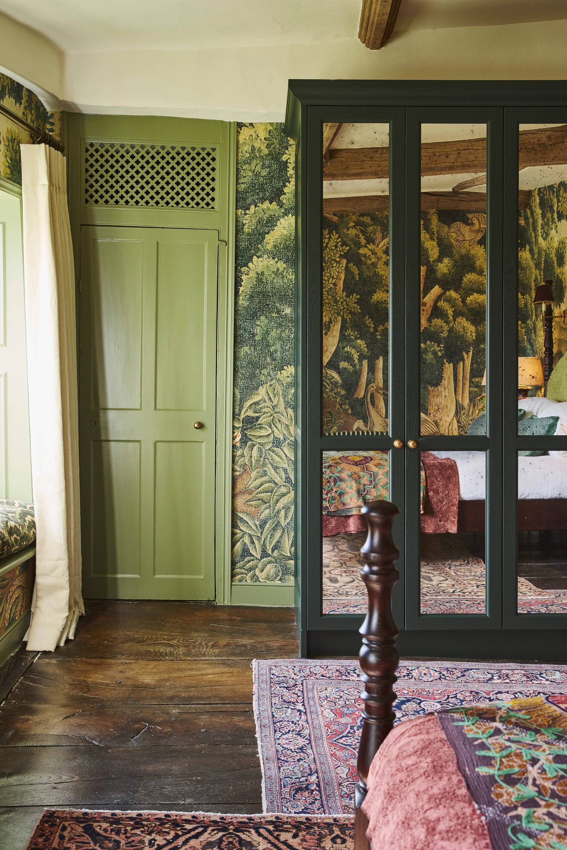 A cozy bedroom with green paneled doors, mirrored wardrobes, wood flooring, patterned rugs, and wallpaper featuring a forest scene. Part of a bed and a window with light curtains are also visible.