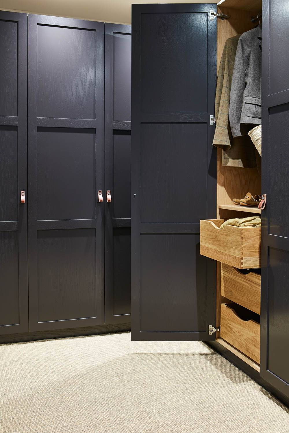 A modern dark wood wardrobe with one door open, revealing wooden drawers, neatly folded clothes, and a hanging jacket inside. The wardrobe stands on a light-colored carpeted floor