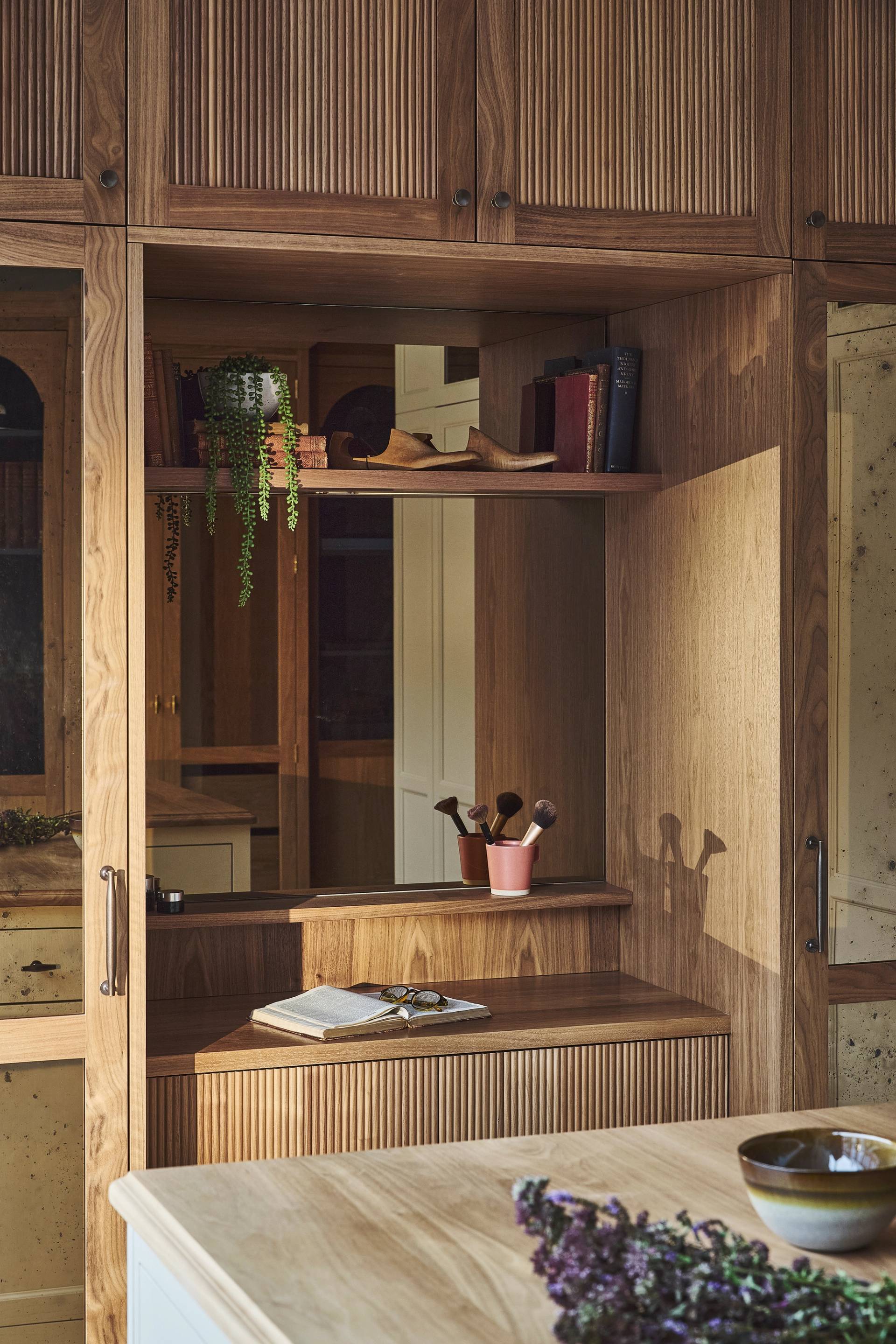 A cozy wooden vanity with shelves, books, plants, and makeup brushes sits next to a large mirror. A checkered ottoman is in front, and sunlight streams in from a window, creating a warm and inviting atmosphere.