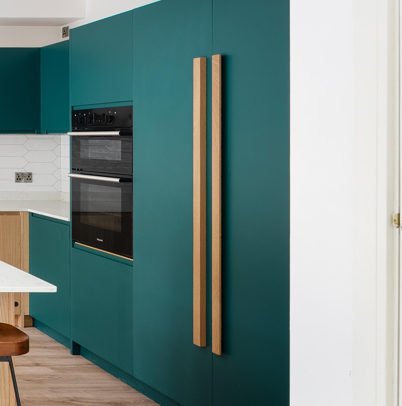 Modern kitchen with teal cabinets, wooden handles, built-in black oven, white hexagonal tile backsplash, and a white countertop. Part of a wooden chair and light wood floor are also visible.