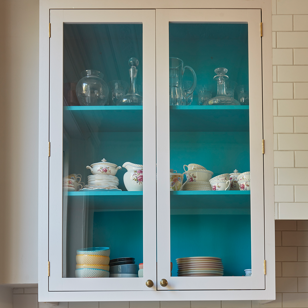 A white kitchen cabinet with glass doors displays glassware, teacups, teapots, and plates on blue shelves. The background features white subway tile.