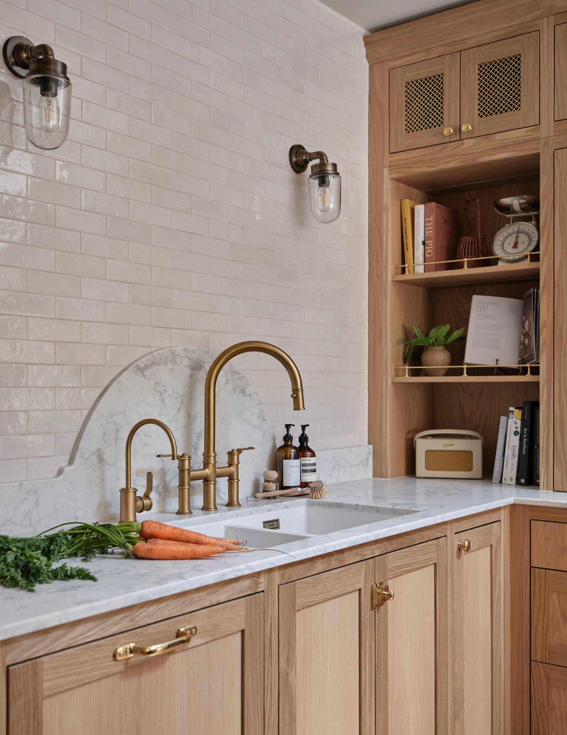 A modern kitchen with light wood cabinets, brass fixtures, a marble countertop and backsplash, carrots on the counter, open shelves with books, a scale, and a cream-colored radio.