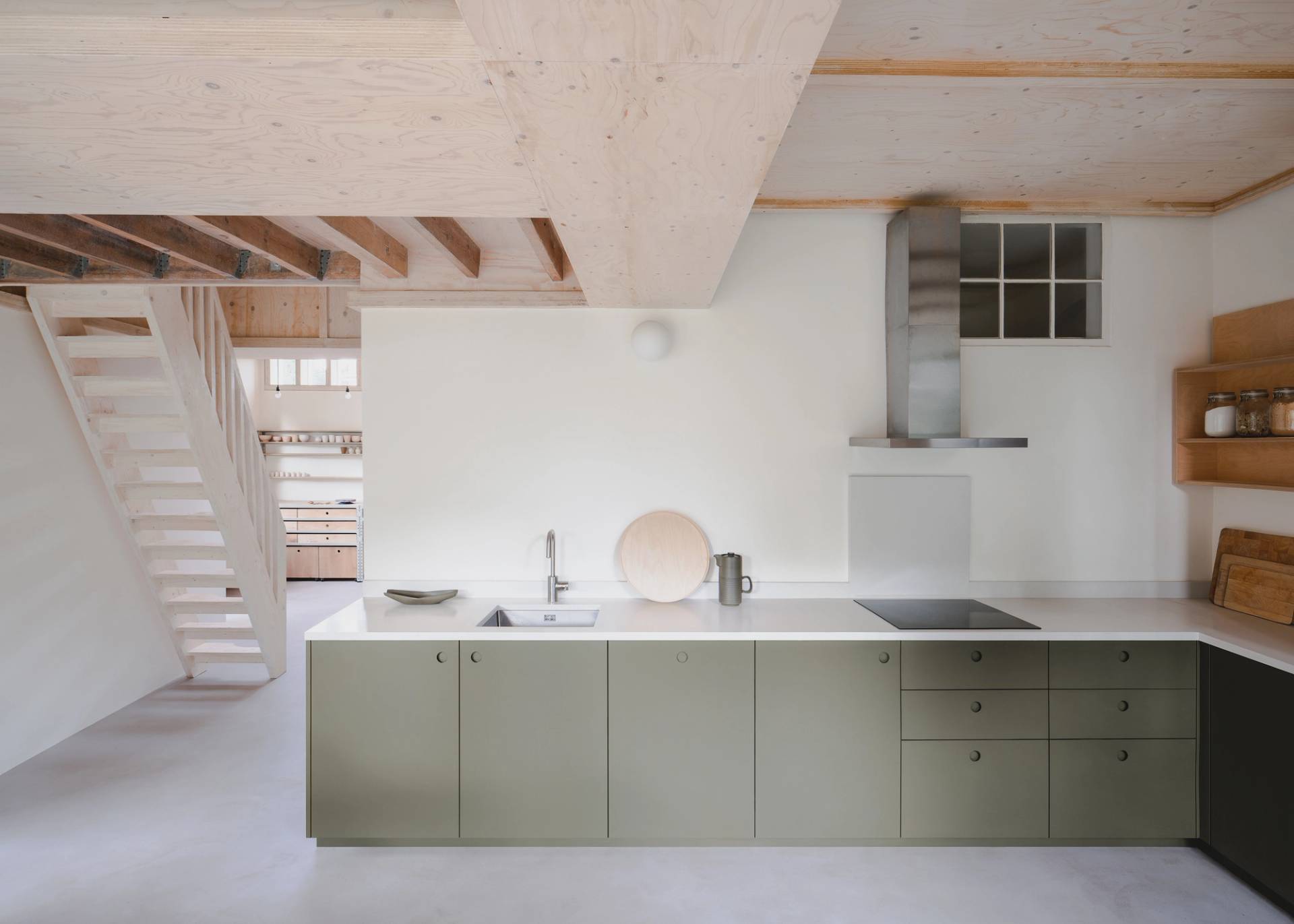 Minimalist kitchen with green cabinets, white countertops, and a sleek modern design. Includes a stovetop, sink, and shelves with jars. Light wood ceiling and open staircase in the background enhance the airy feel
