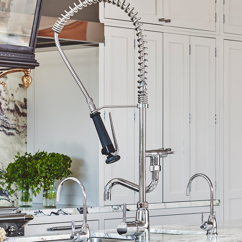 A modern kitchen sink with a tall, spring-coiled pull-down faucet, two smaller faucets, marble countertop, and white cabinetry in the background. A plant sits near the backsplash.