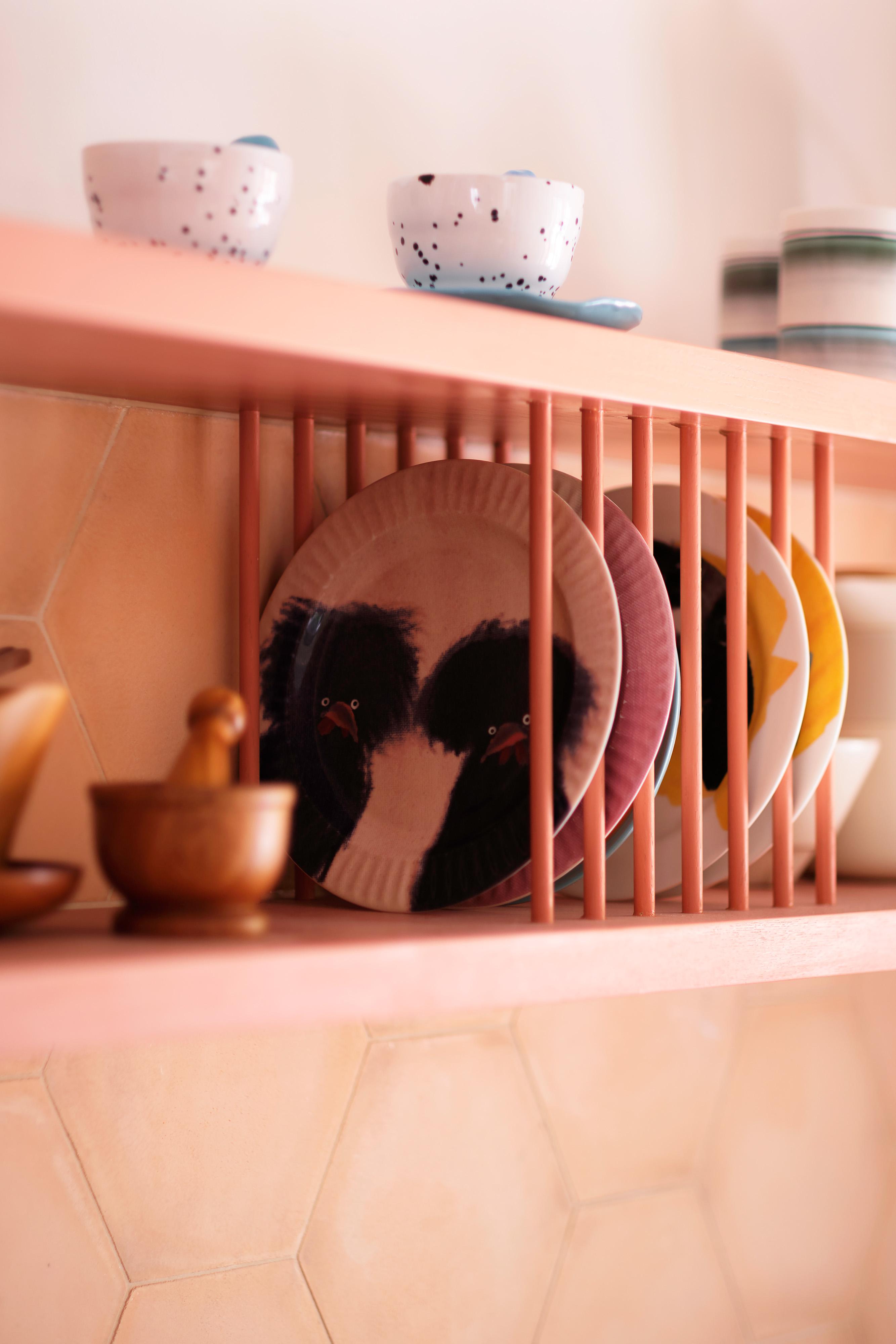 A close-up of a pink dish rack holding colorful plates and bowls. On top, speckled cups and saucers are stacked. The background features a light hexagonal tile backsplash.