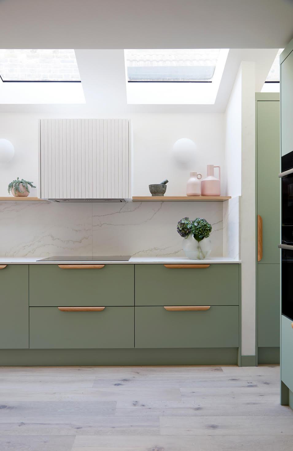 Modern kitchen with sage green cabinets and wooden handles. Marble backsplash and light wood flooring. Skylights above illuminate the space. Decor includes pink vases and small plants on a wooden shelf