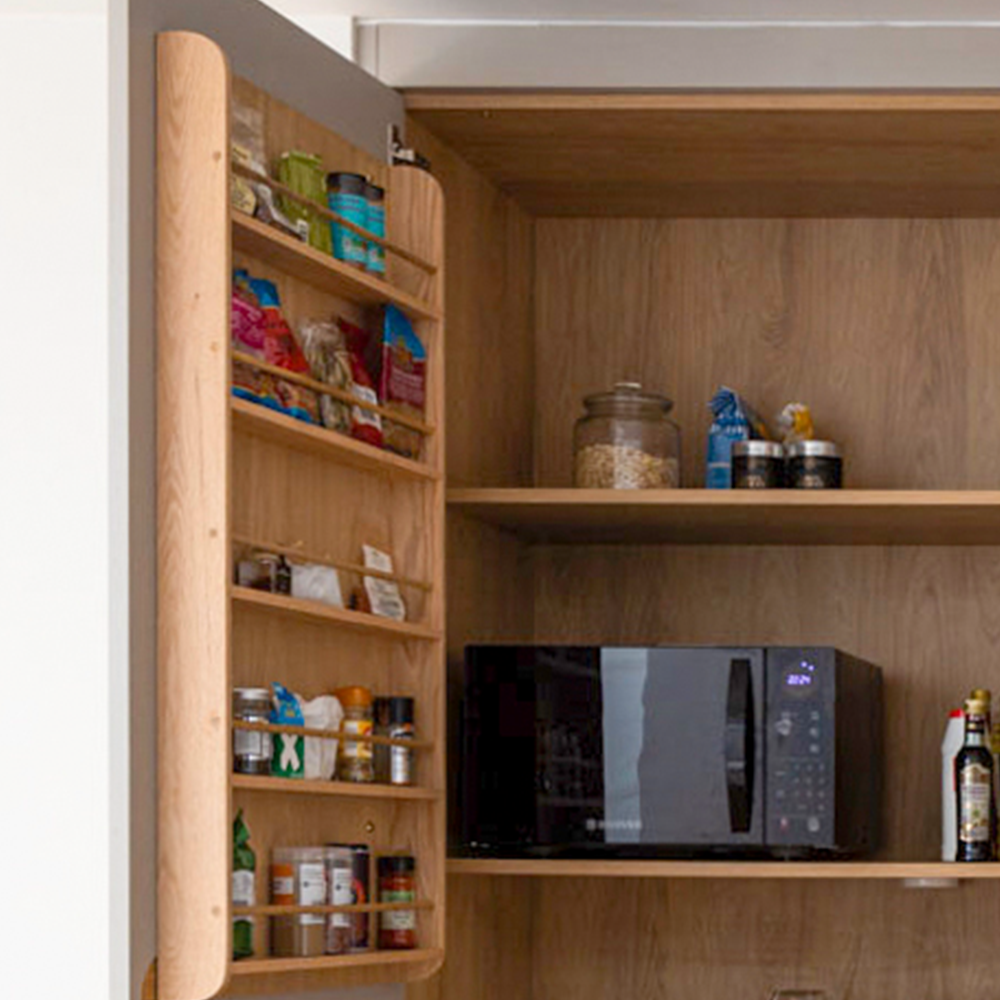 An open kitchen cabinet reveals shelves with a microwave, jars, and containers. The inside of the cabinet door has racks holding spices and small bottles. The background features wood paneling.