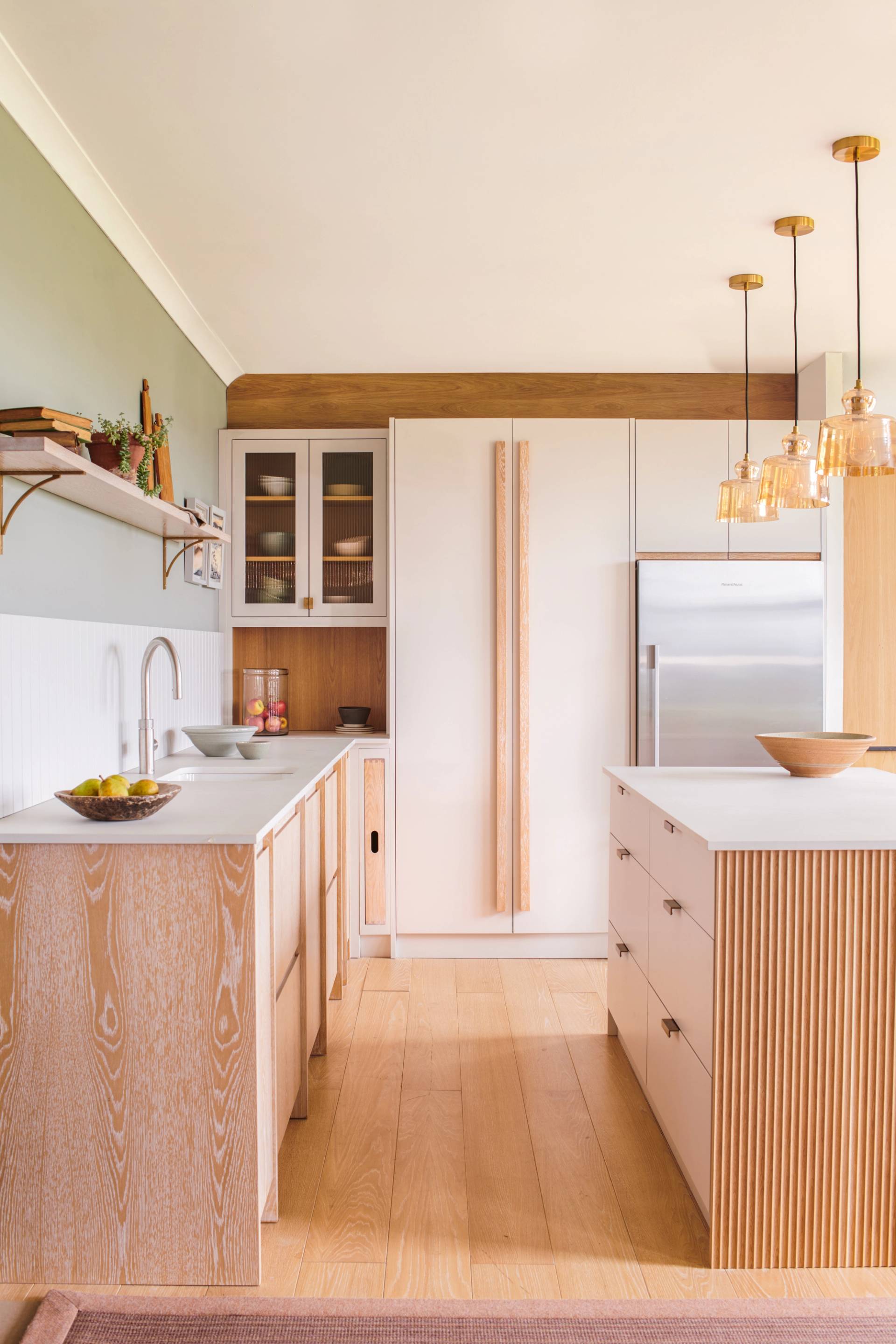 A modern kitchen with light wood cabinets, a large island with vertical wood paneling, a pink stool, pendant lights, white countertops, open shelving, and a stainless steel refrigerator.