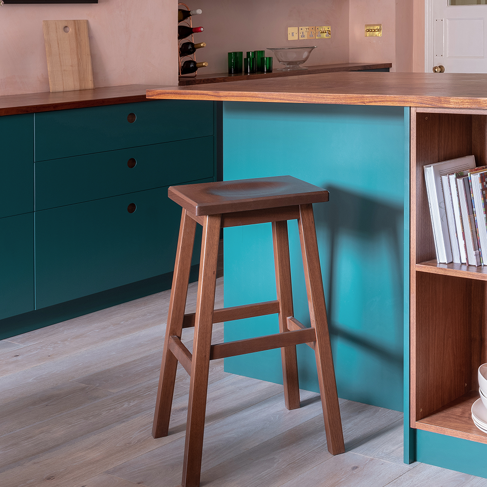 A wooden stool stands by a kitchen island with teal cabinets, open shelving, and books. The kitchen has wood countertops, light-colored walls, and light wood flooring. Wine bottles are visible on a side shelf.