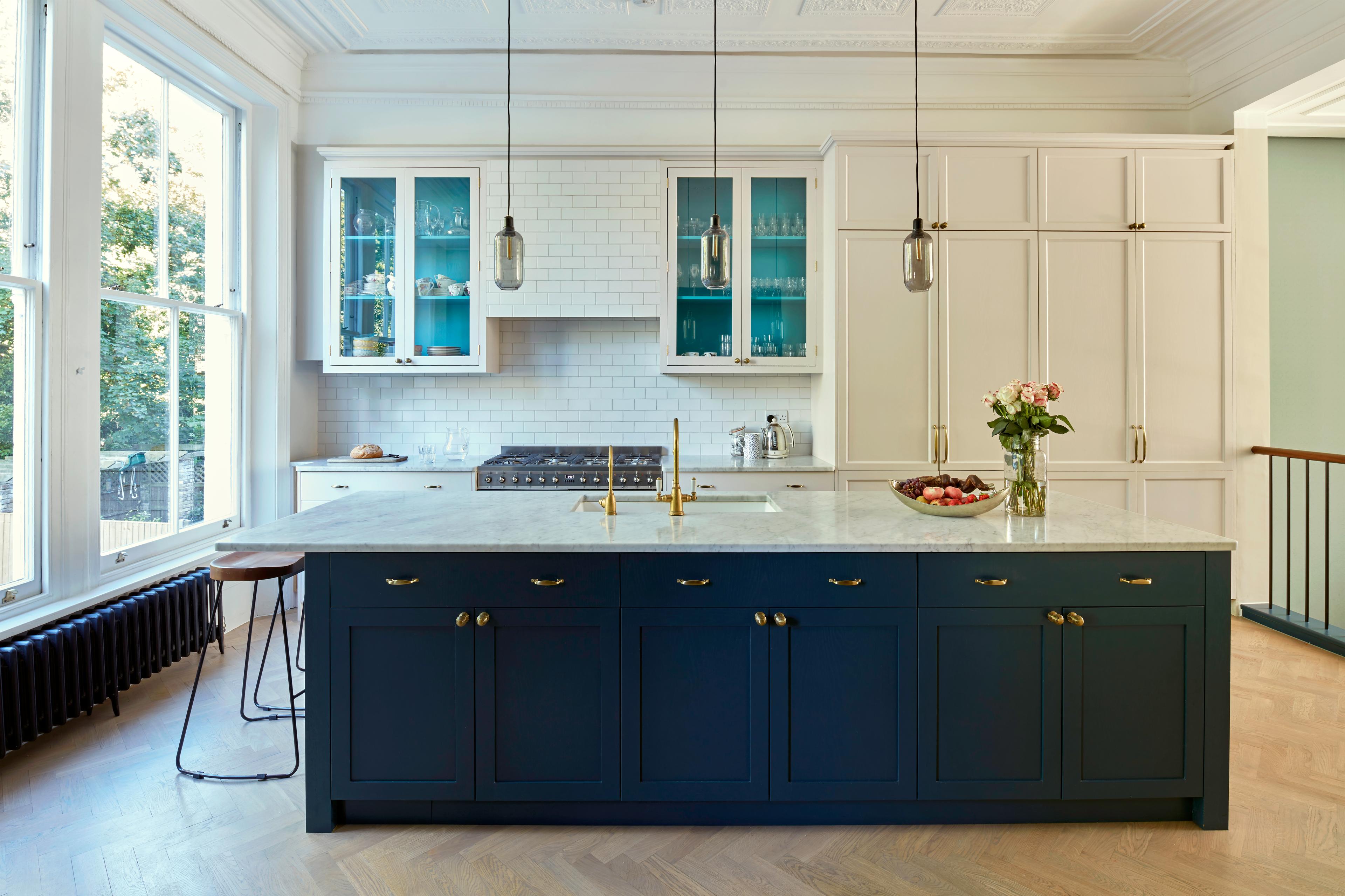 A modern kitchen with a navy blue island featuring a marble countertop and gold handles. Three pendant lights hang above. White cabinets and subway tiles line the walls, with large windows letting in natural light. Flowers and a fruit bowl are on the island.
