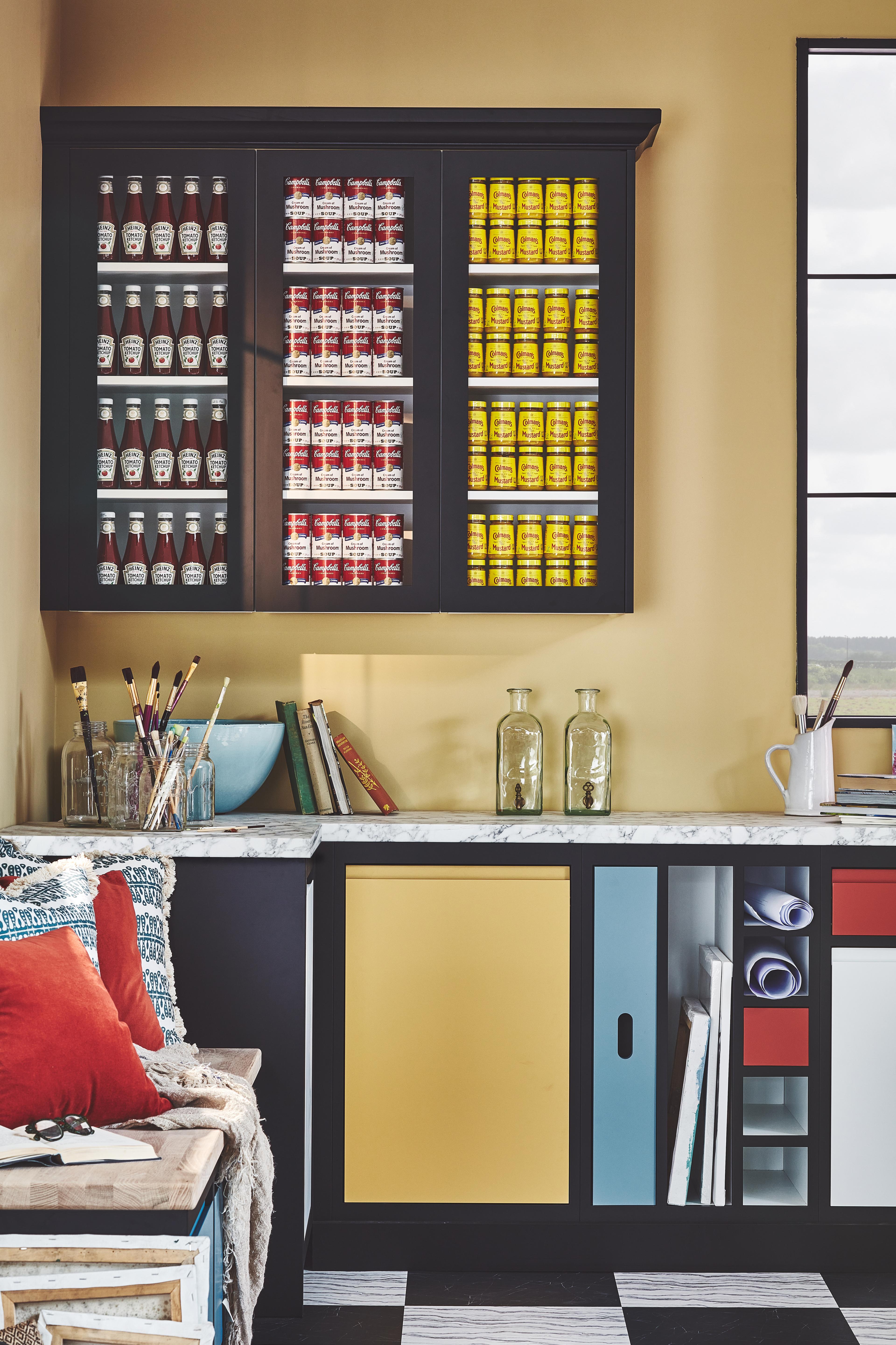 A close-up of a colorful art studio workspace featuring a black glass-front cabinet filled with rows of ketchup bottles, soup cans, and mustard jars.