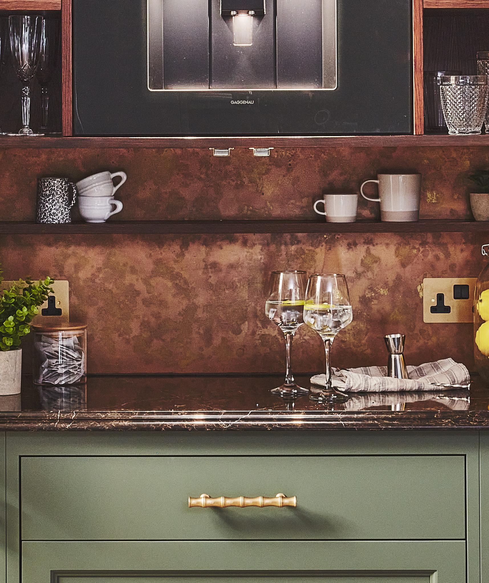 A kitchen counter with a green drawer and gold handle, two glasses with drinks, a napkin, lemons, green plant, mugs and cups on shelves, and a built-in coffee machine above.