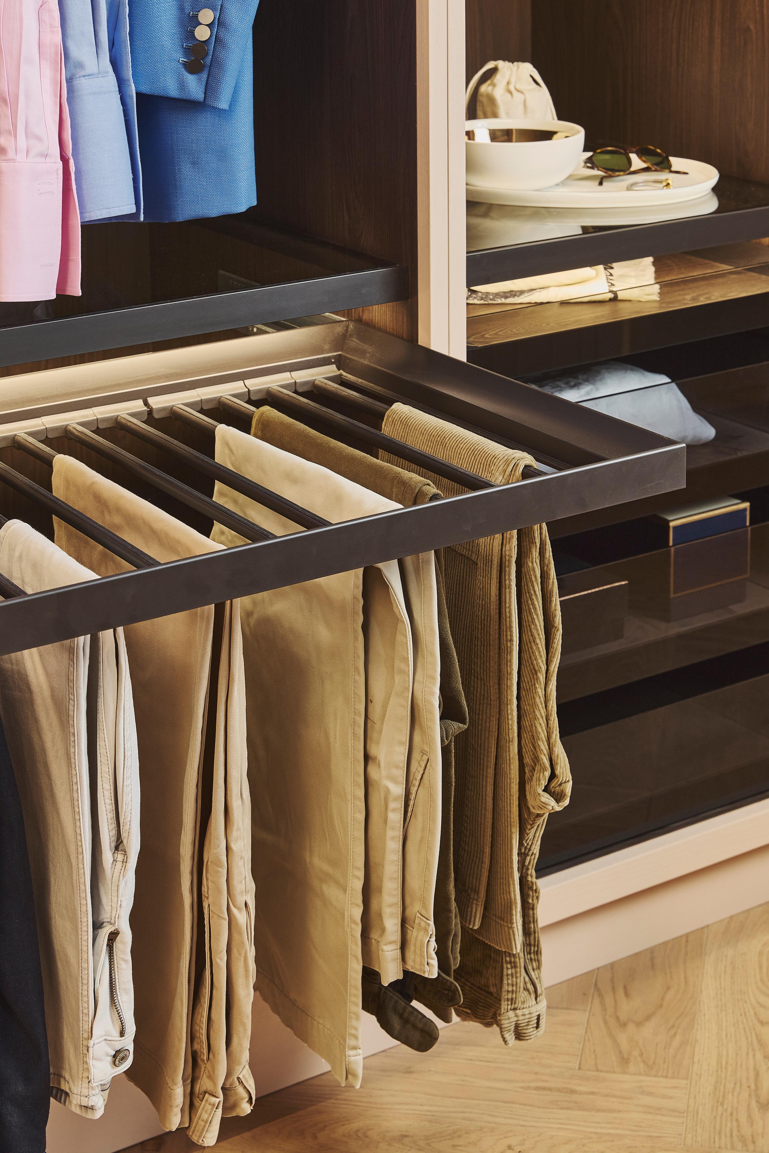 A modern closet with sliding drawers, featuring a pull-out fabric laundry hamper containing clothes, neatly arranged shirts and trousers, and shelves with bowls and sunglasses.