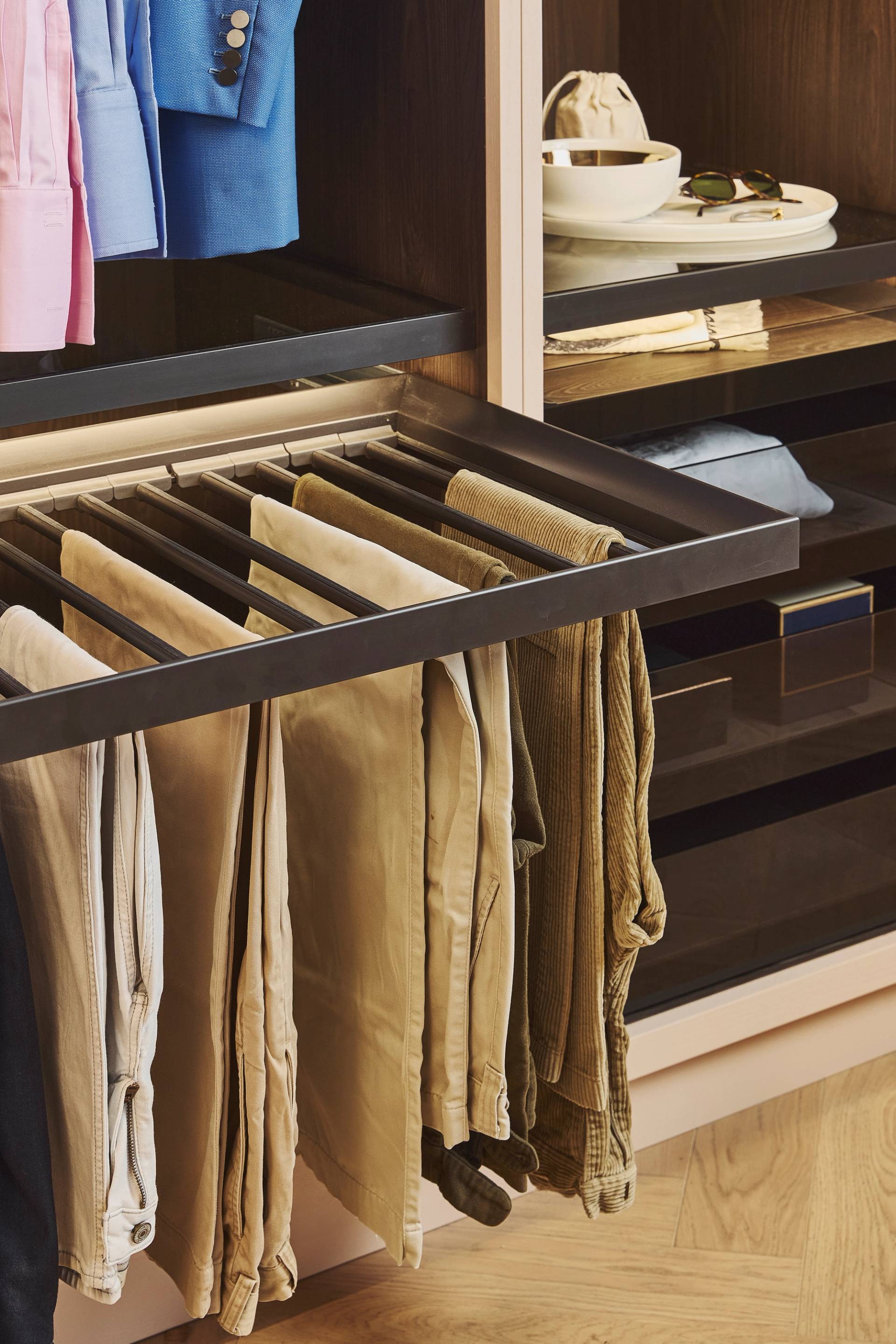 A modern closet with sliding drawers, featuring a pull-out fabric laundry hamper containing clothes, neatly arranged shirts and trousers, and shelves with bowls and sunglasses.