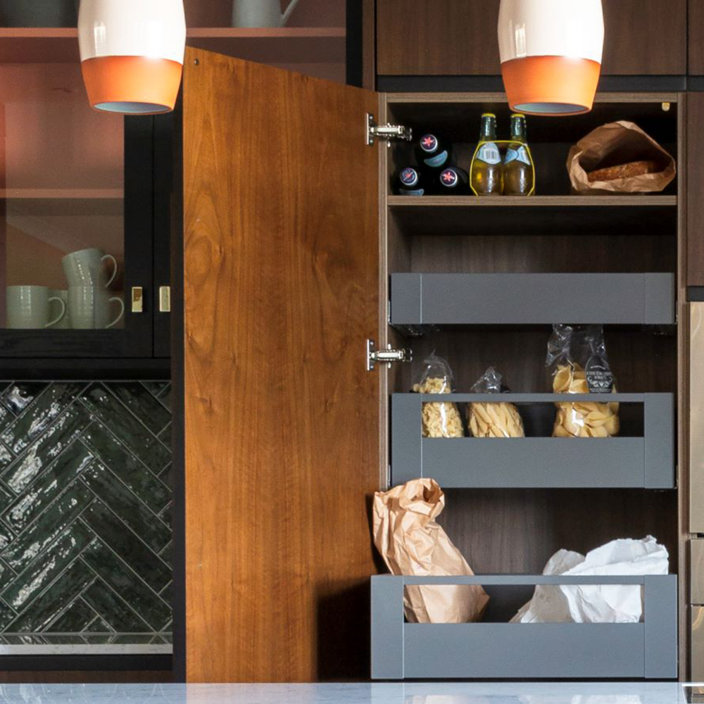 A modern kitchen with dark wood cabinets, some open to reveal shelves stocked with bottled drinks, jars, and groceries in paper bags. White mugs and glassware are seen behind glass doors, and pendant lights hang overhead.