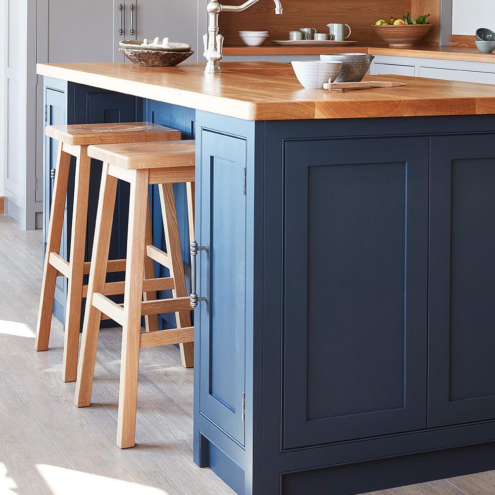 A modern kitchen island with a wooden countertop, navy blue cabinets, and two light wood bar stools. The background shows a bowl, dishes, and other kitchen items on the counter.