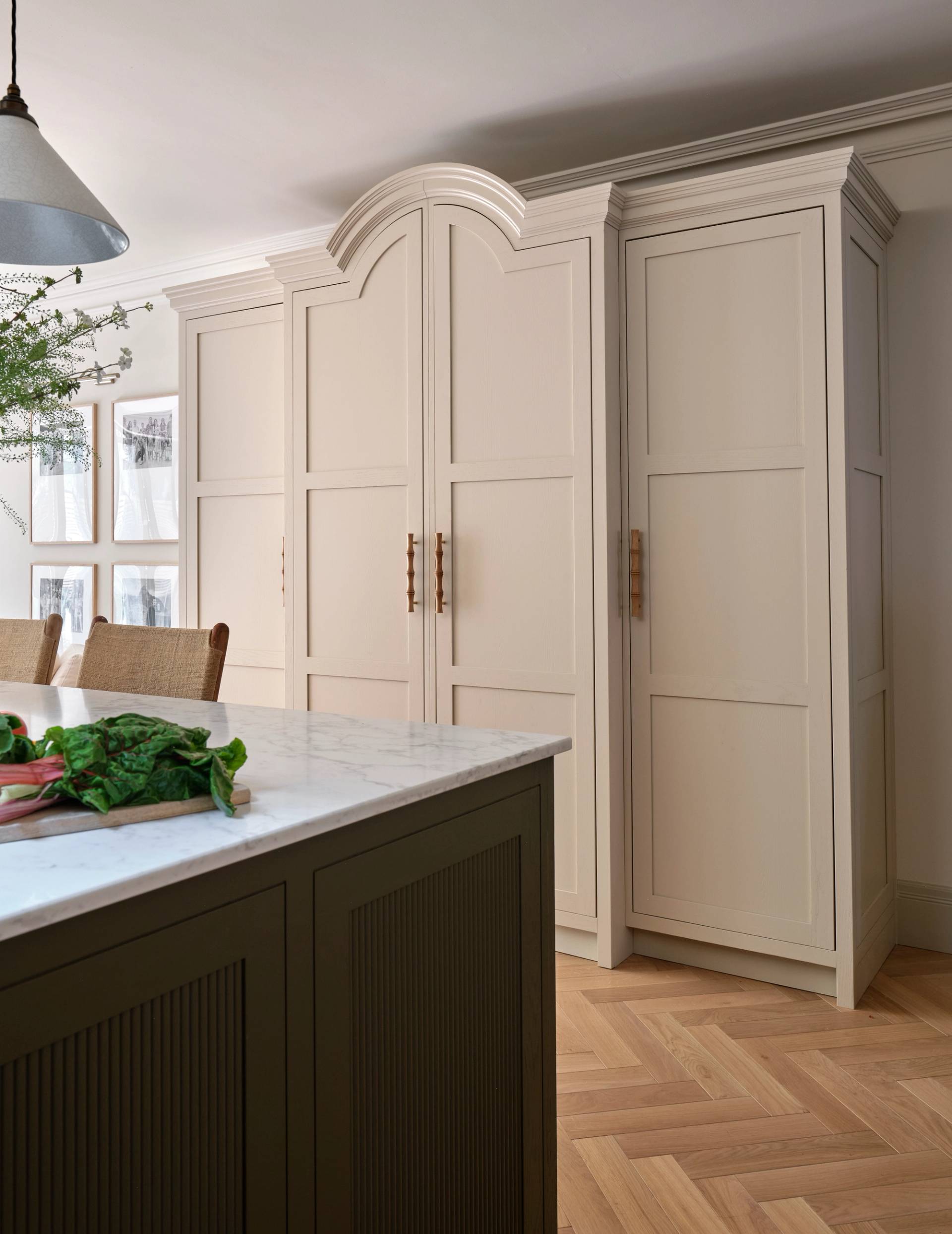 Large cream-colored kitchen cabinets with panel doors and brass handles stand against the wall. An island with a marble countertop and dark base is in the foreground. The floor features a light herringbone wood pattern.